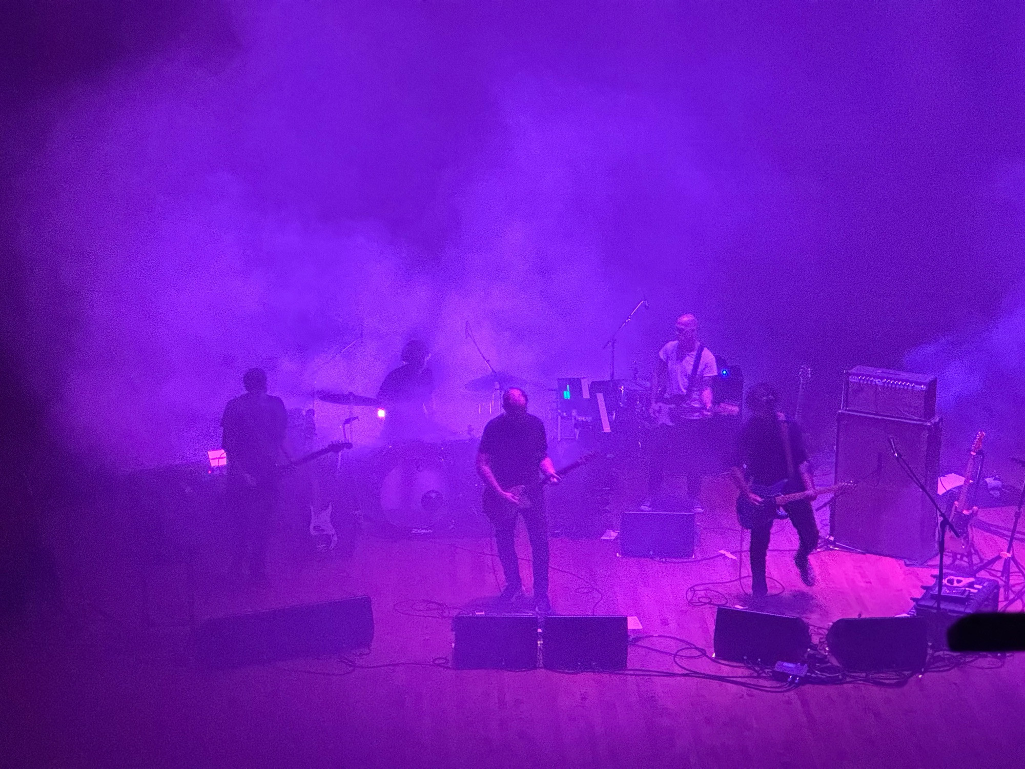 five men on stage, mostly obscured by smoke and lit with purple lights from multiple low angles