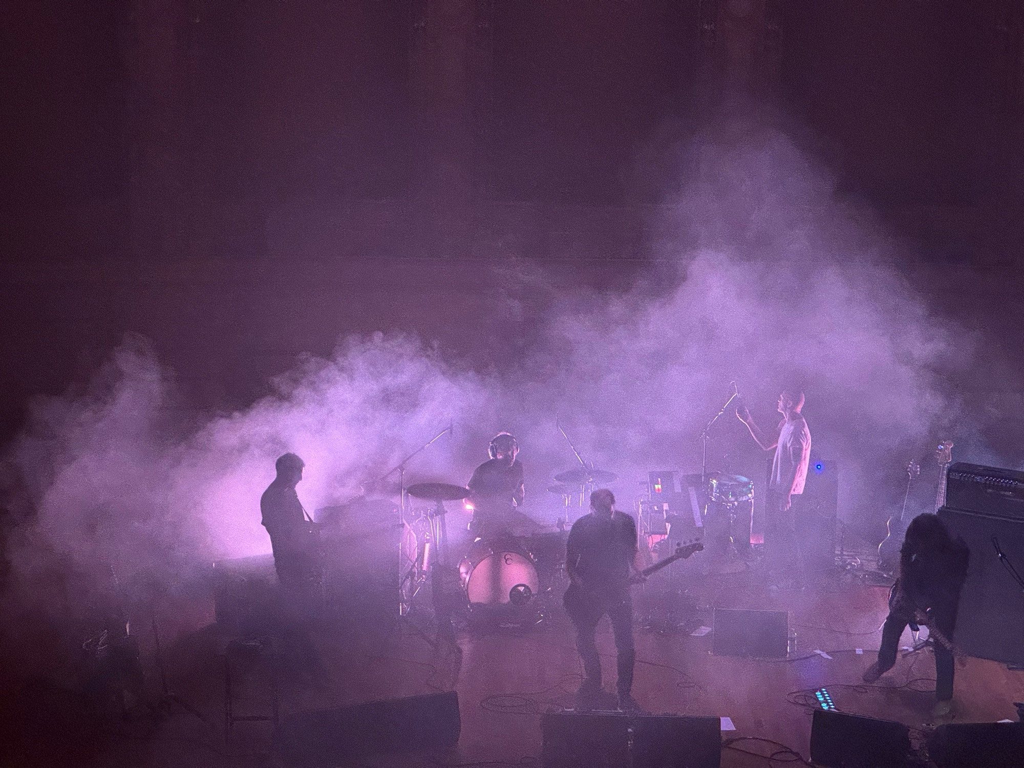 five men on stage, mostly obscured by smoke and lit with white lights from multiple low angles