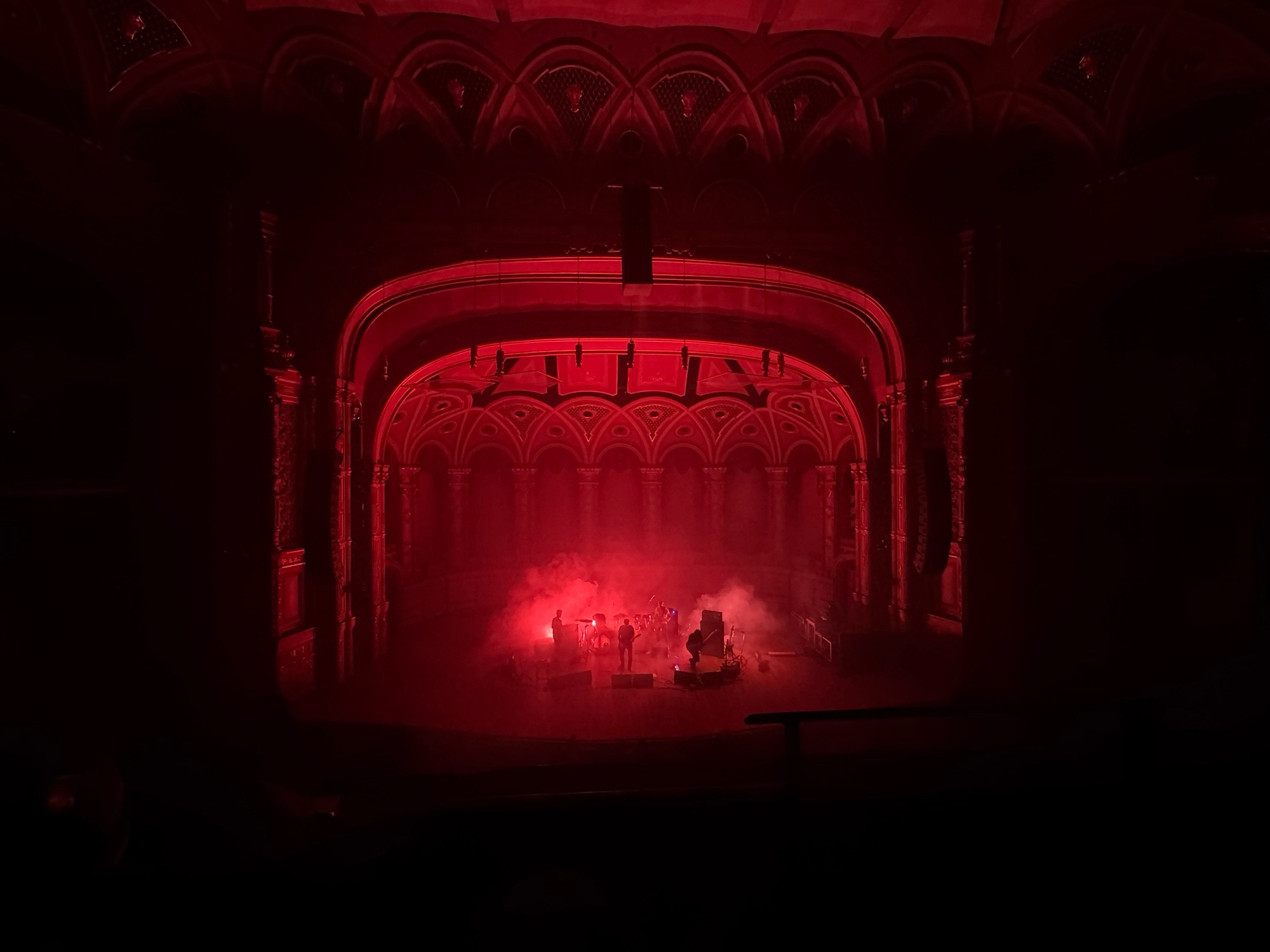 a red-lot faraway shot of a band on stage, figures barely visible behind smoke, and the arch and decorations of the venue visible