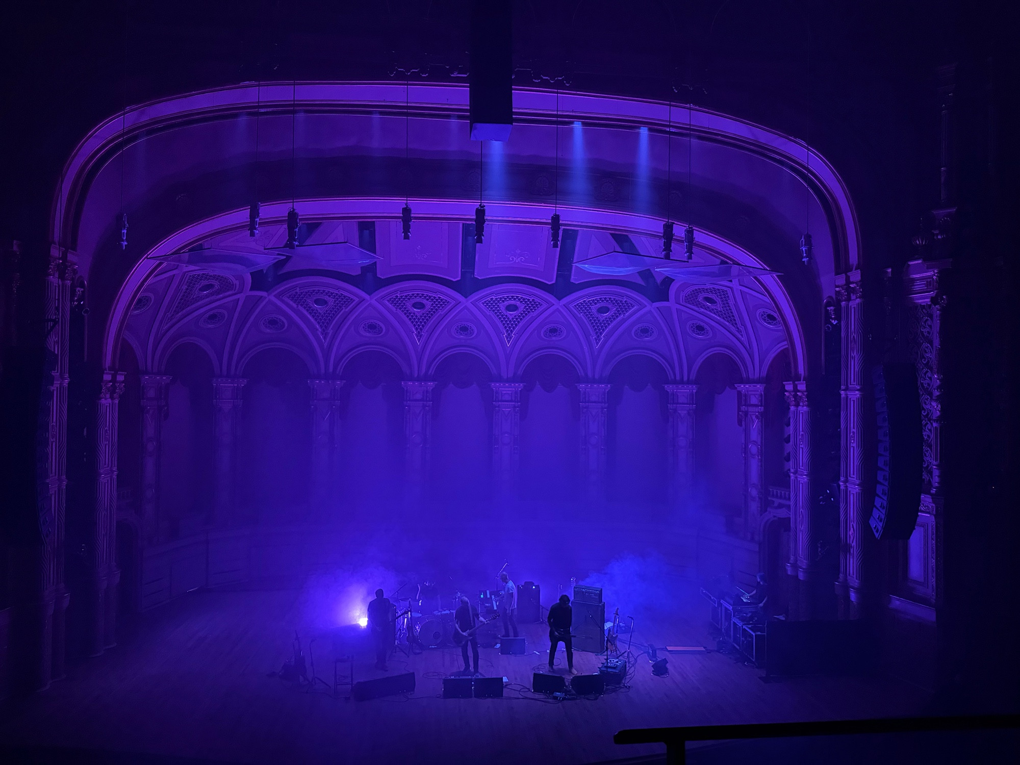 five men on stage, mostly obscured by smoke and lit with purple lights from multiple low angles. the angle is wide enough to show the whole stage and art deco elements behind the band