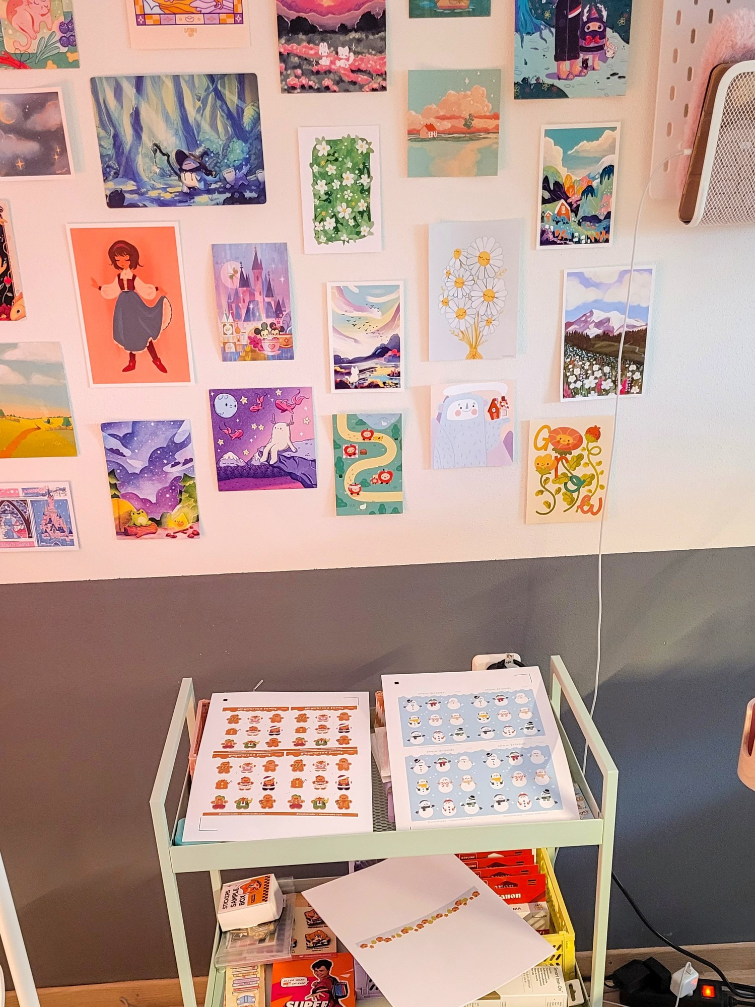 A picture of an art wall filled with different beautiful art prints of different artists. Underneath the prints on the wall is a cart, or art cart, filled with tools, samples and prints that still need to be cut into stickers. 