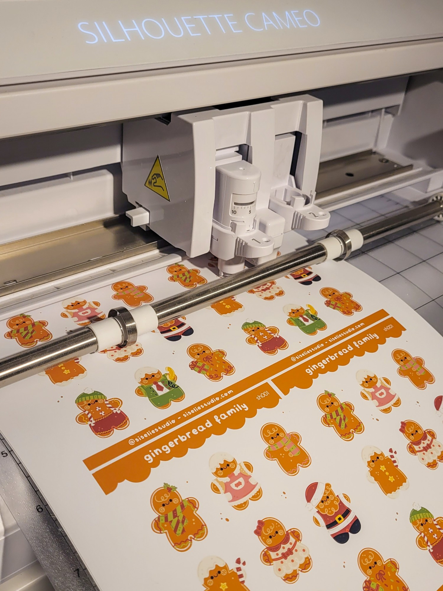 A picture of a cutting machine cutting 'gingerbread family' sticker sheets filled with cute gingerbread characters. Each one with a different cute and Christmas or seasonal outfit. 