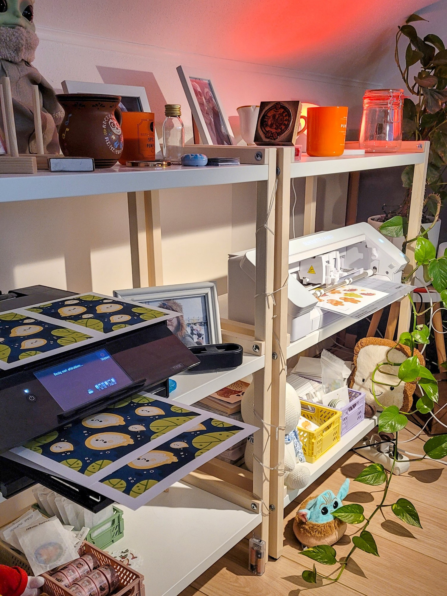 A picture of a messy studio space with cozy orange light and a plant growing between the trinkets. Among all the plushies, candles, products and cozy chaos are a printer and cutting machine hard at work. 