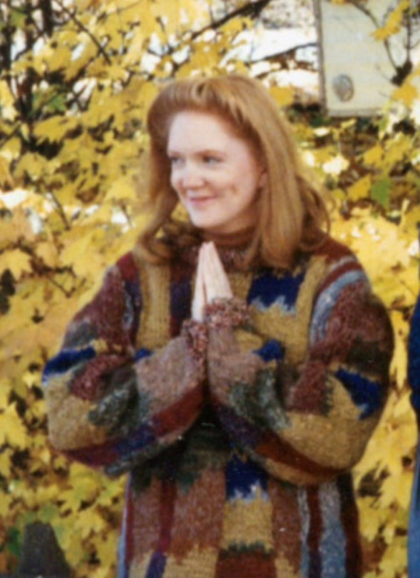 Photo of a hottie redhead wearing a multi-colored sweater and with her hands in the prayer position. She stands in front of a tree with yellowish leaves, indicating autumn.