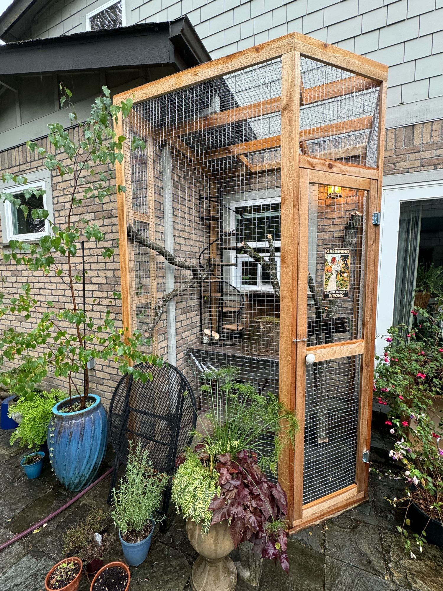 A catio built against the side of a house with a spiral plant stand, a table, a couple of wooden crates, and several tree branches for climbing.