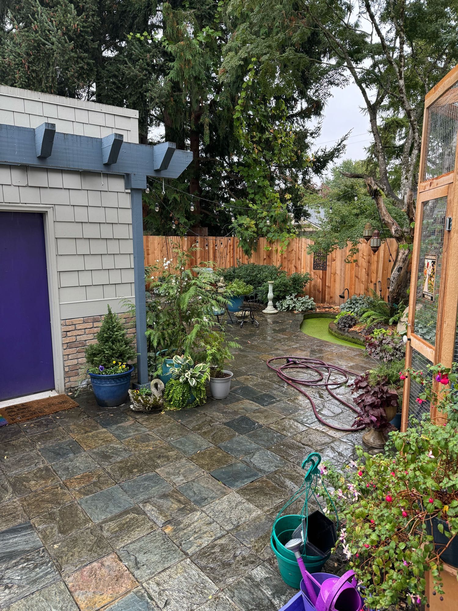 Image of a hardscaped backyard with a garage on the left, plants in pots on the ground, and a wood fence.