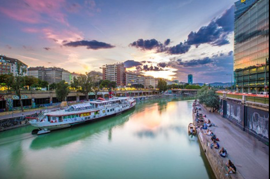 Zu sehen ist die Donau samt des Donaukanals. Auf der Donau ist ein Schiff. Am Rand des Kanals sitzen die Menschen auf dem Boden. 