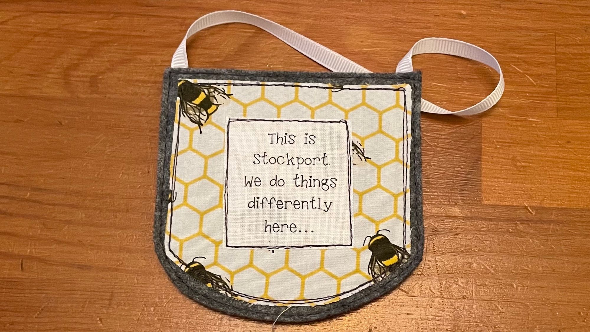 A small fabric patch on a hanging ribbon. It has a design featuring a yellow honeycomb pattern and bees, and the text:
This is Stockport.
We do things differently here…