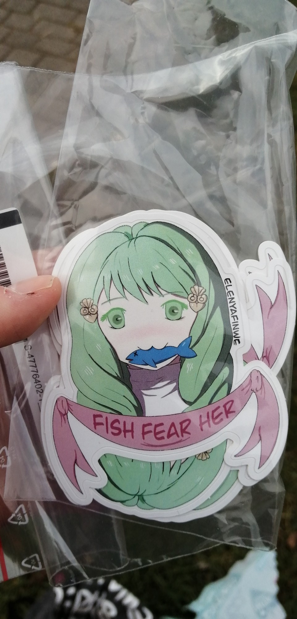 I'm holding a bunch of stickers of Flayn making a :3 face and having a fish in her mouth. A pink banner under her portrait reads "Fish Fear Her".