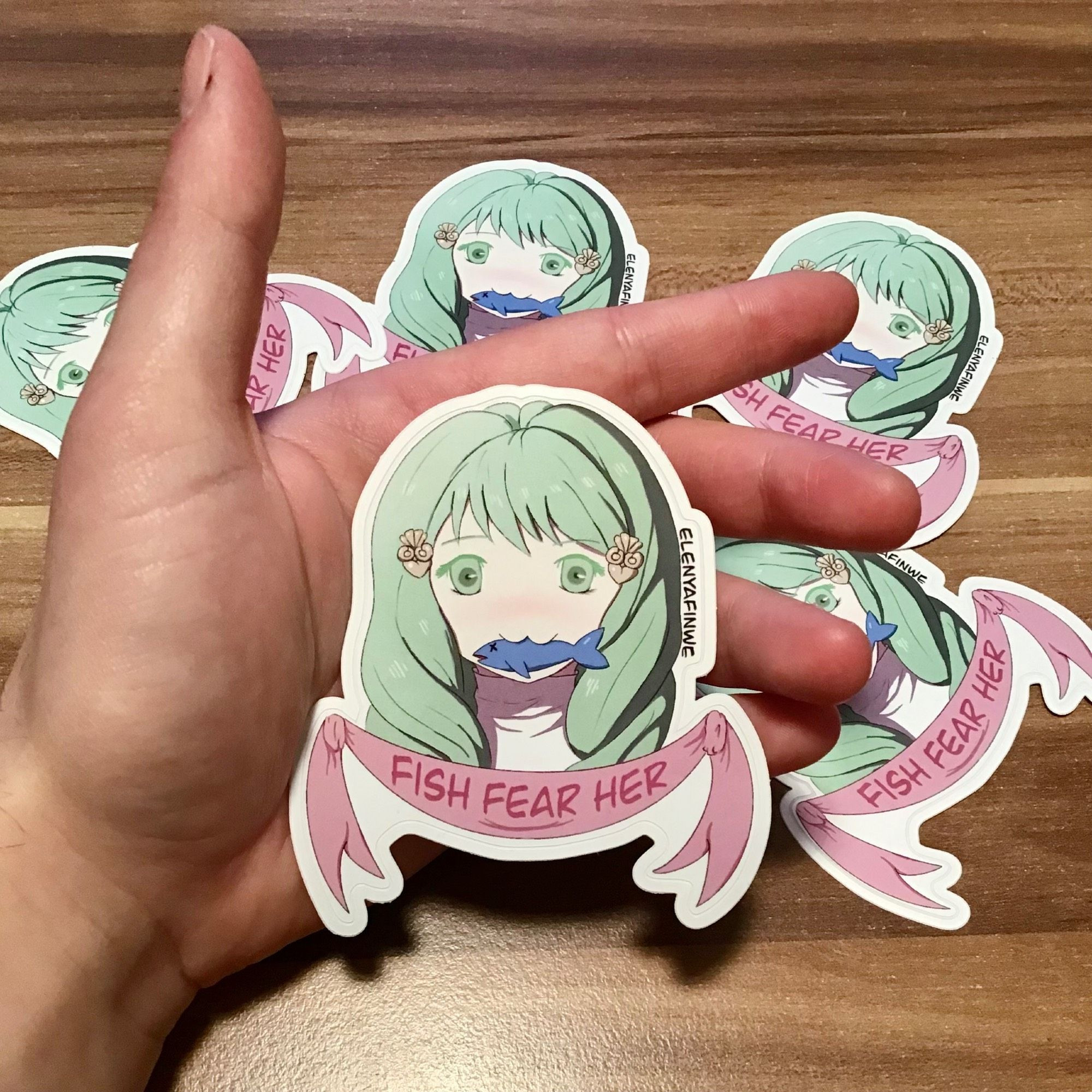 I'm holding a Flayn sticker. She makes a :3 face and has fish in her mouth. A pink banner under her portrait reads Fish Fear Her