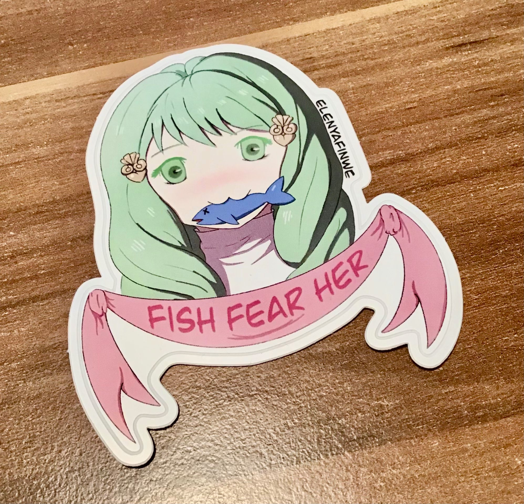 Sticker of Flayn making a :3 face with a fish in her mouth. A banner under her portrait reads „Fish Fear Her“