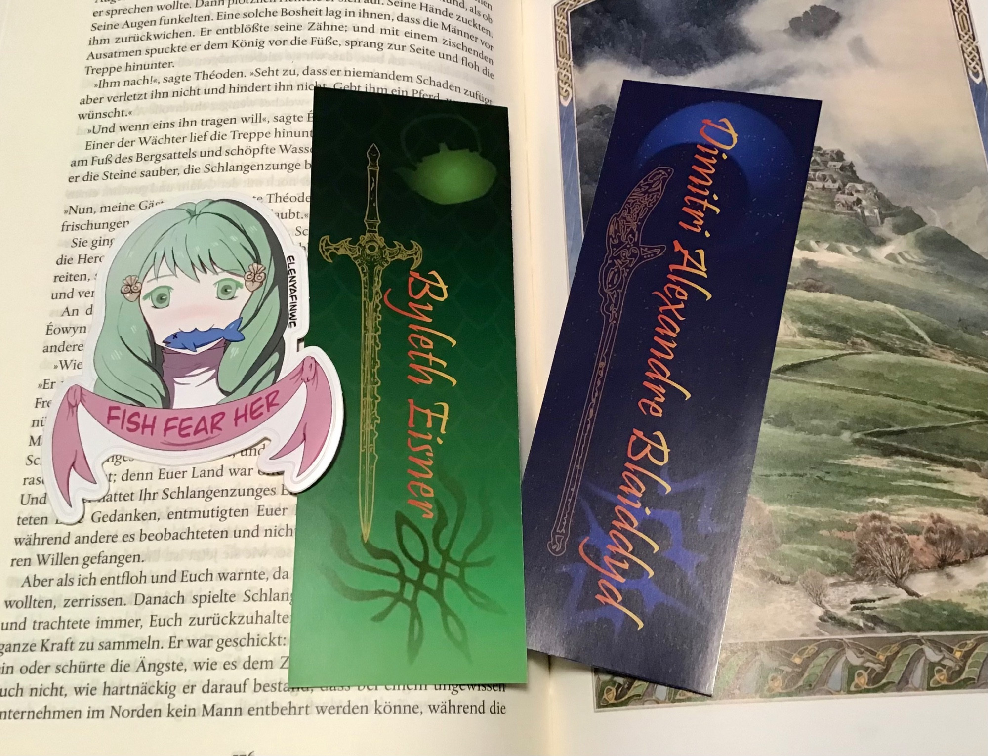 The sticker together with two bookmarks on an open book. One bookmark is green gradient with The Sword of the Creator, a tea kettle and the Crest of Flames and a text reading Byleth Eisner. The other bookmark is dark blue with the Blaiddyd crest, a crestent moon, Areadhbar and a text that reads Dimitri Alexandre Blaiddyd