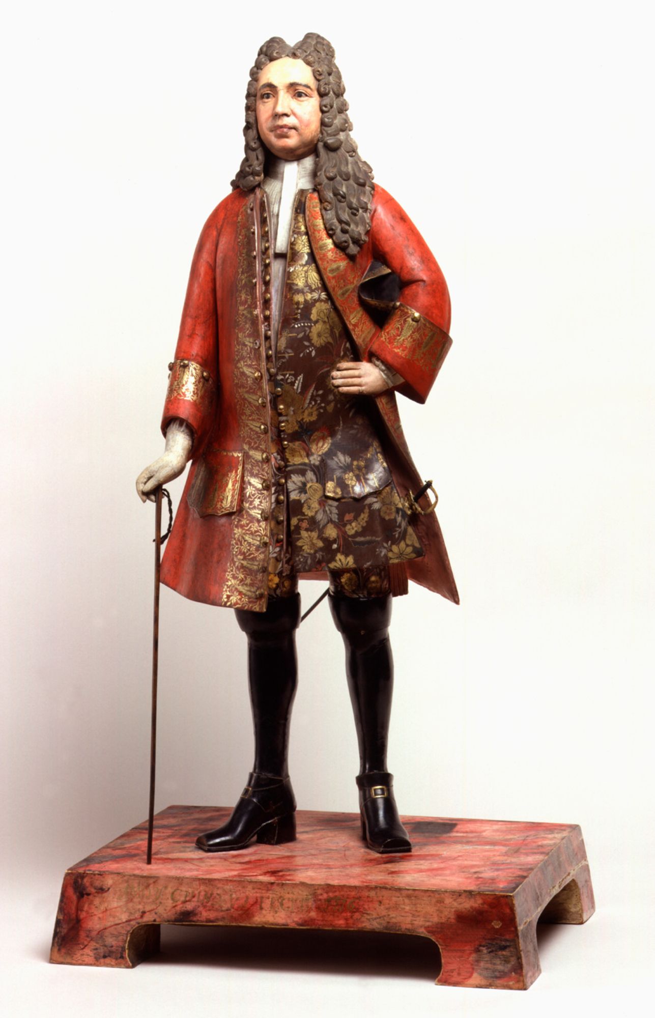 A sculpted clay figurine of a man in a red coat and ornate doublet, holding his hat.