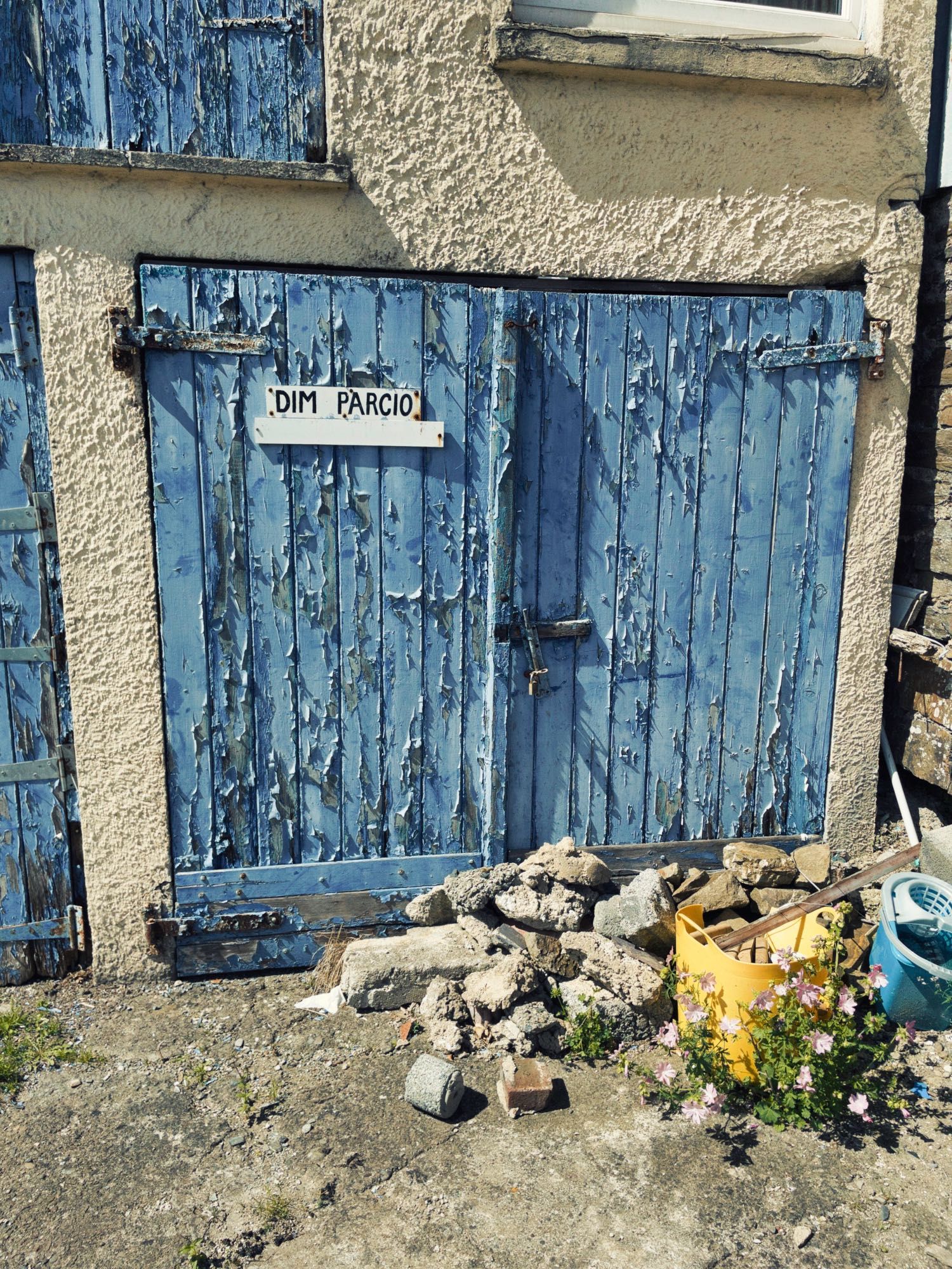 Old garage door with flaking blue paint and ‘dim parcio’ sign. There is a pile of rubble and other rubbish obstructing the door which doesn’t appear to have been opened for a long time. Llandysul, Wales. (Dim Parcio is Welsh for no parking).