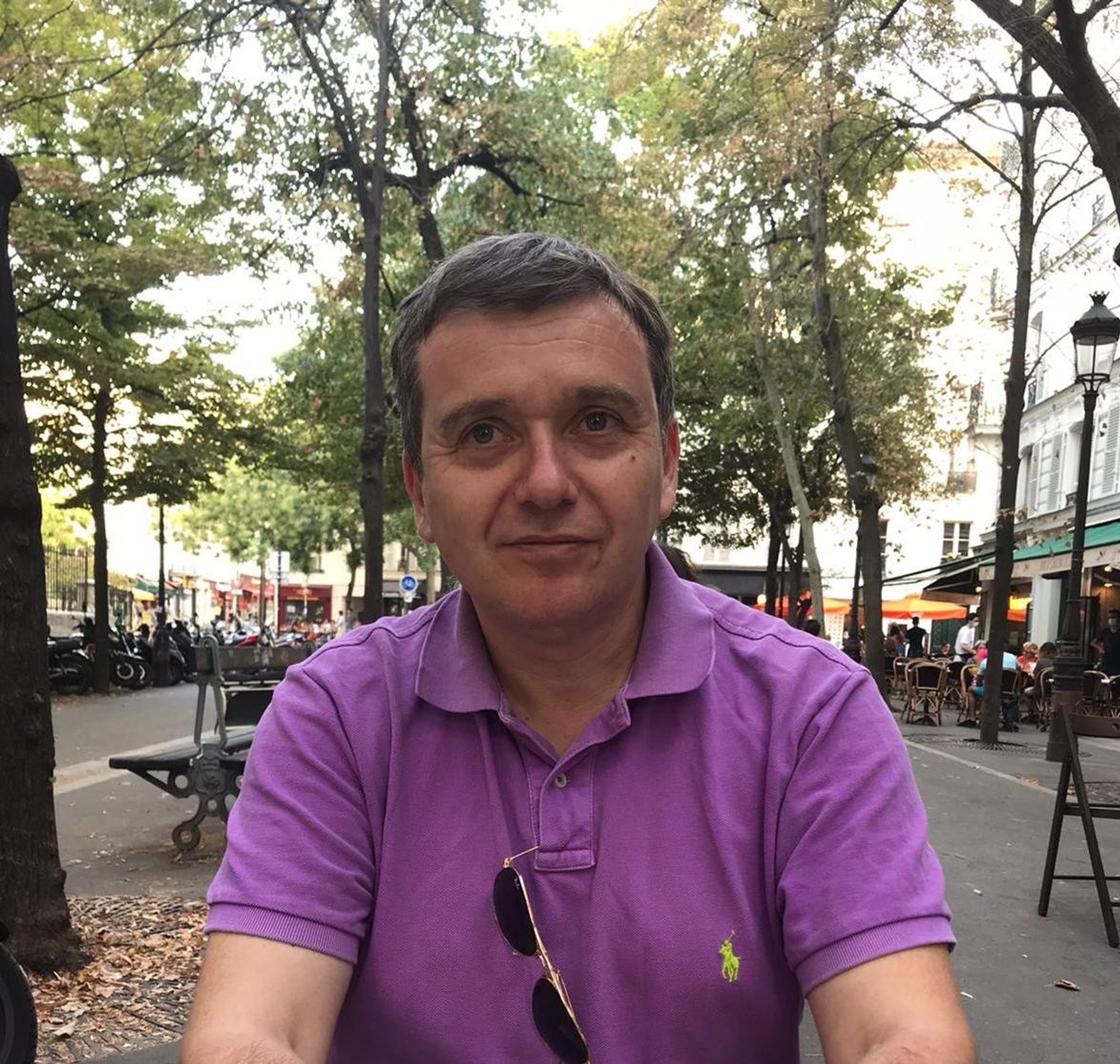 Dr. Damian Catani, University of London.  Photo of a man wearing a purple shirt, on a picturesque city street, under green trees.