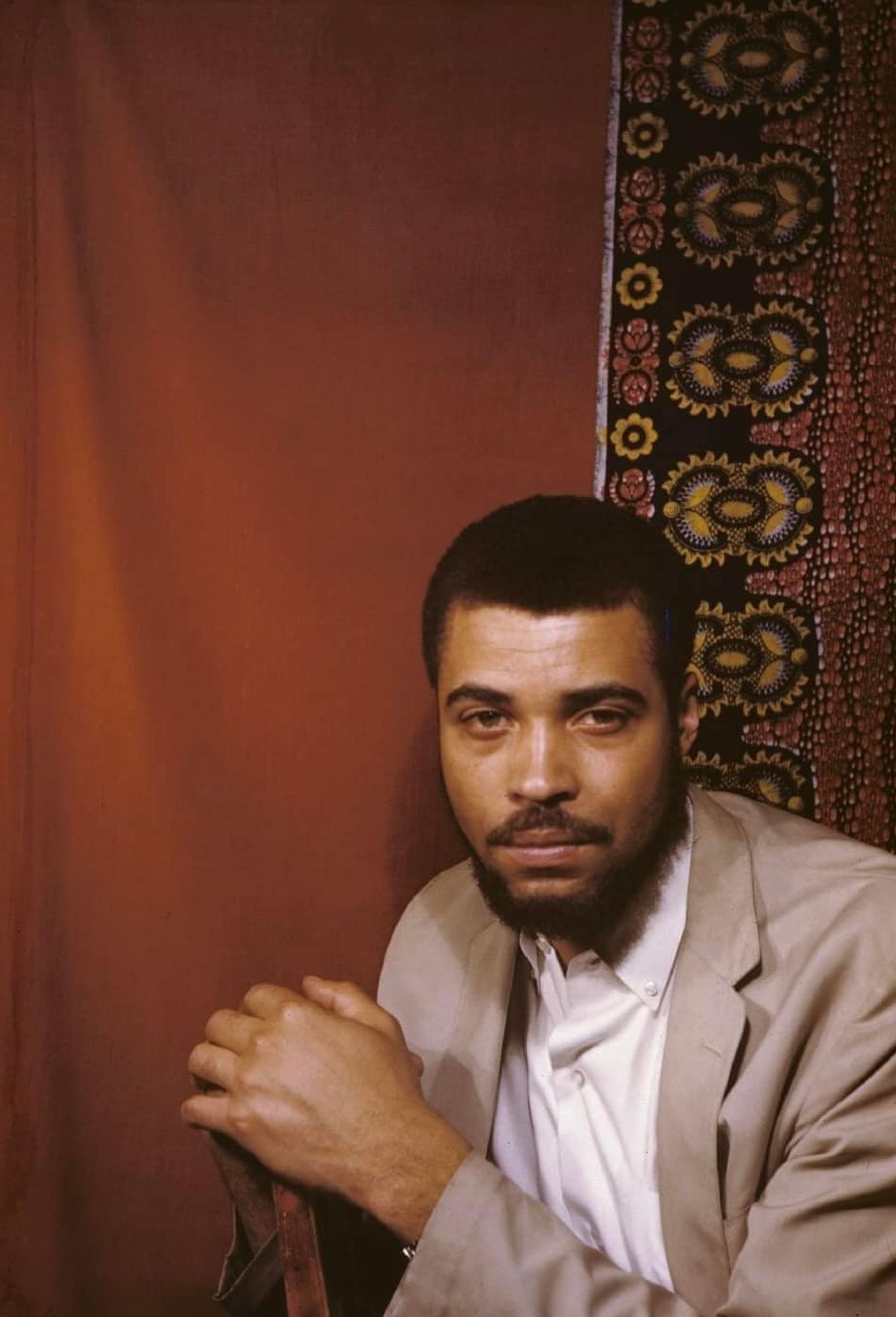 Color photo of young JEJ in a brown suit sitting against a chair back looking at camera, with a red textile backdrop