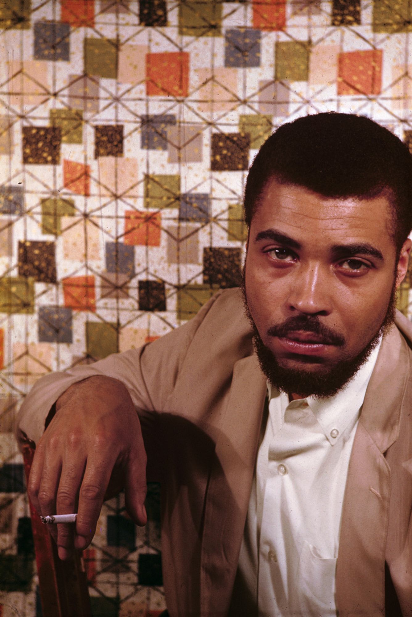 Color photo of young JEJ in a brown suit sitting with a cigarette in his hand, staring mournfully at the camera