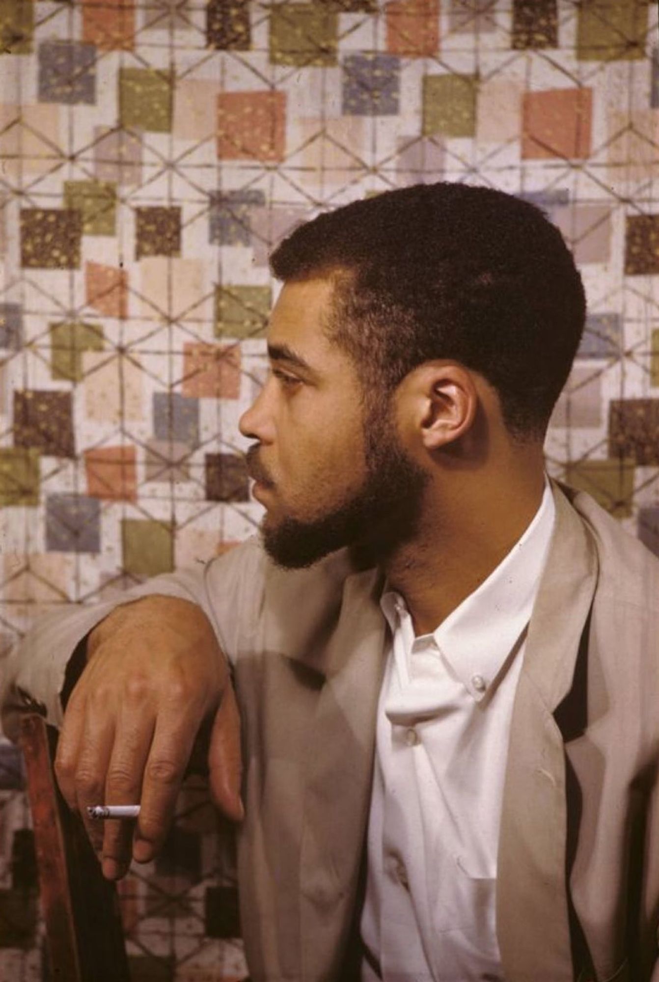 Color photo of young JEJ in a brown suit sitting against a chair back with a cigarette in hand, his face in profile