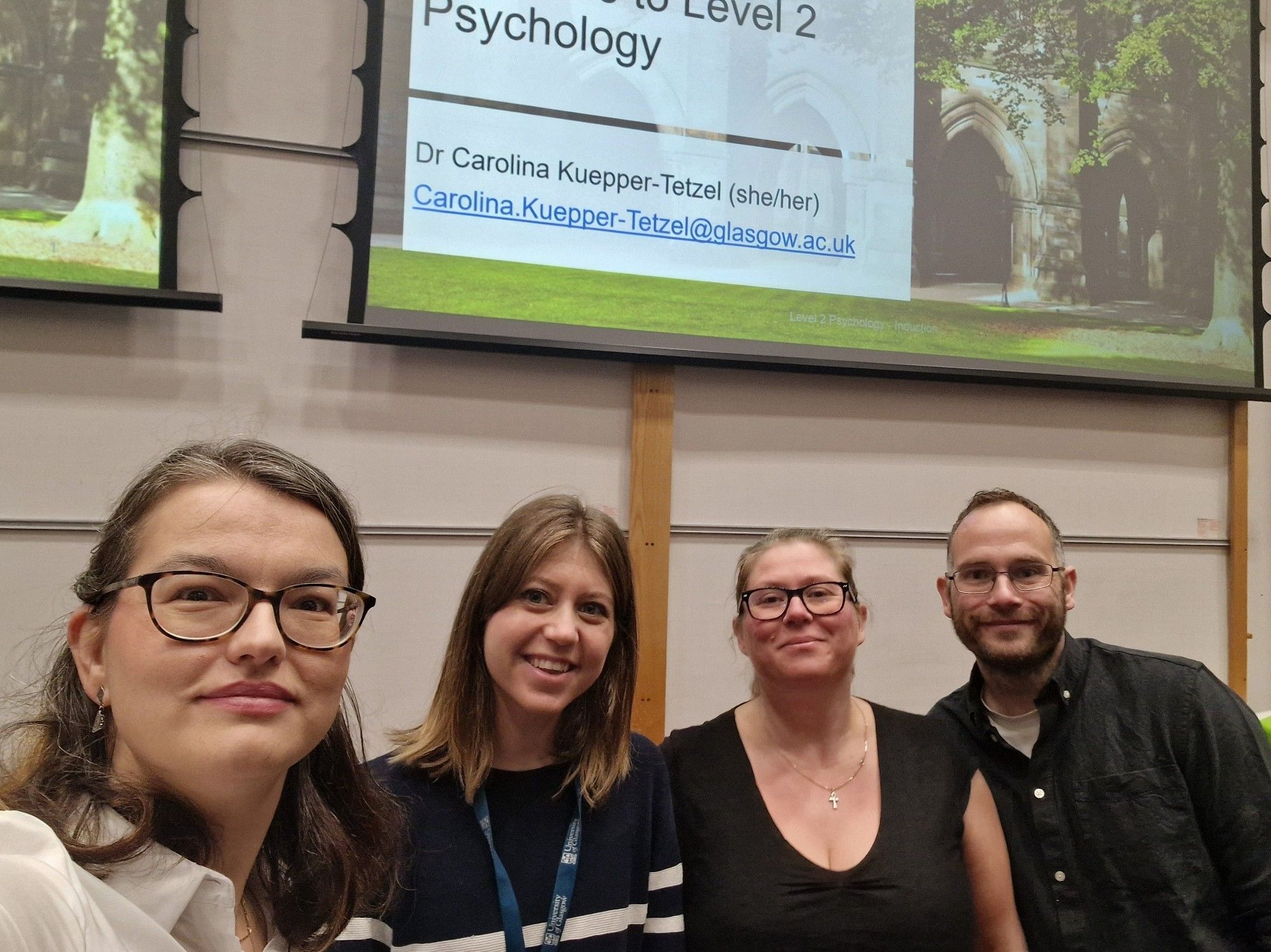 A selfie with four people on it. Left to right: Carolina Kuepper-Tetzel, Jéromine Payot, Gaby Mahrholz, and Jamie Murray. In the background is the intro slide partially displayed. The picture was taken in the lecture hall.