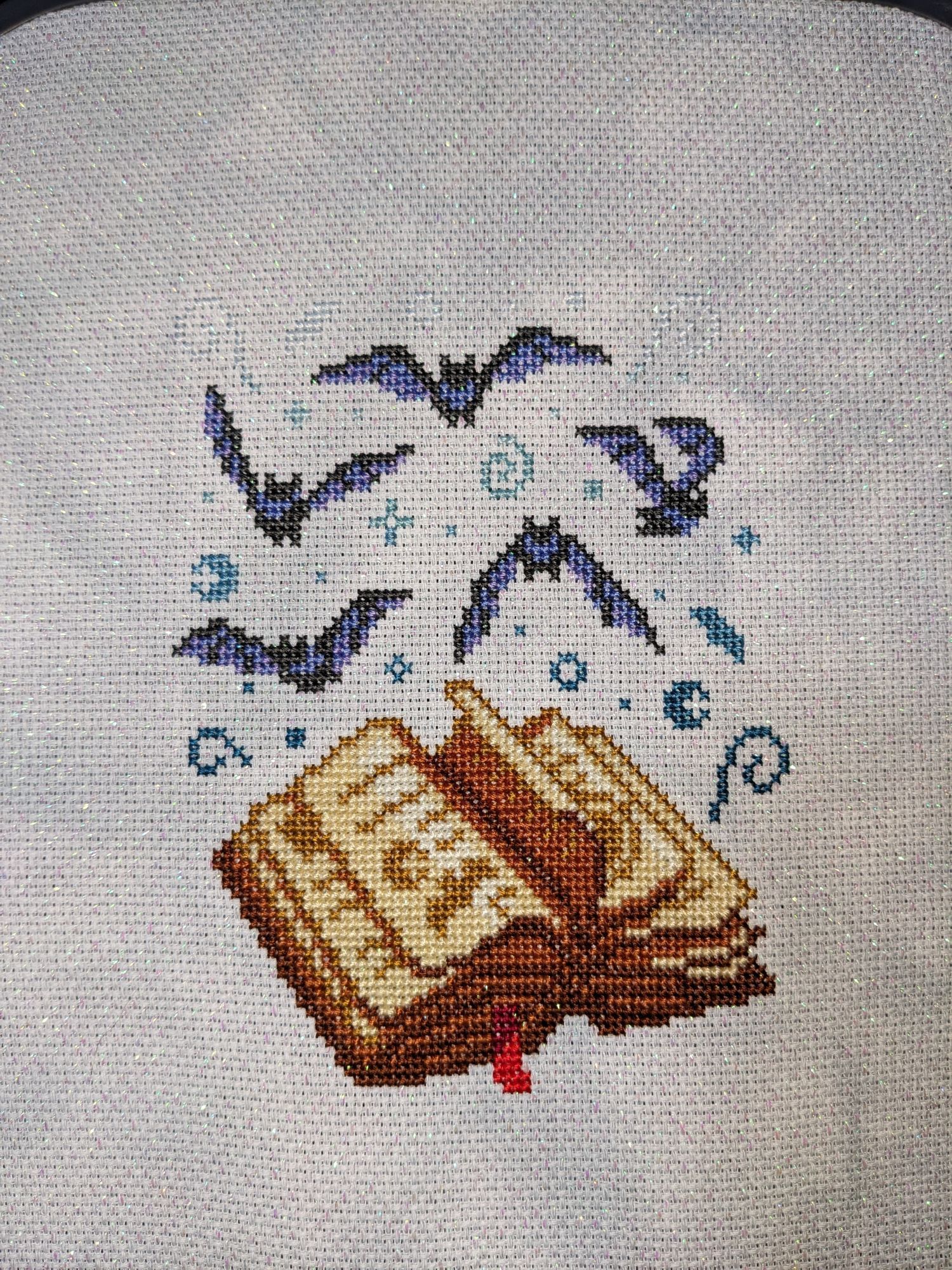 Finished cross stitch piece on opalescent primarily white fabric of an open book with bats flying out of it.