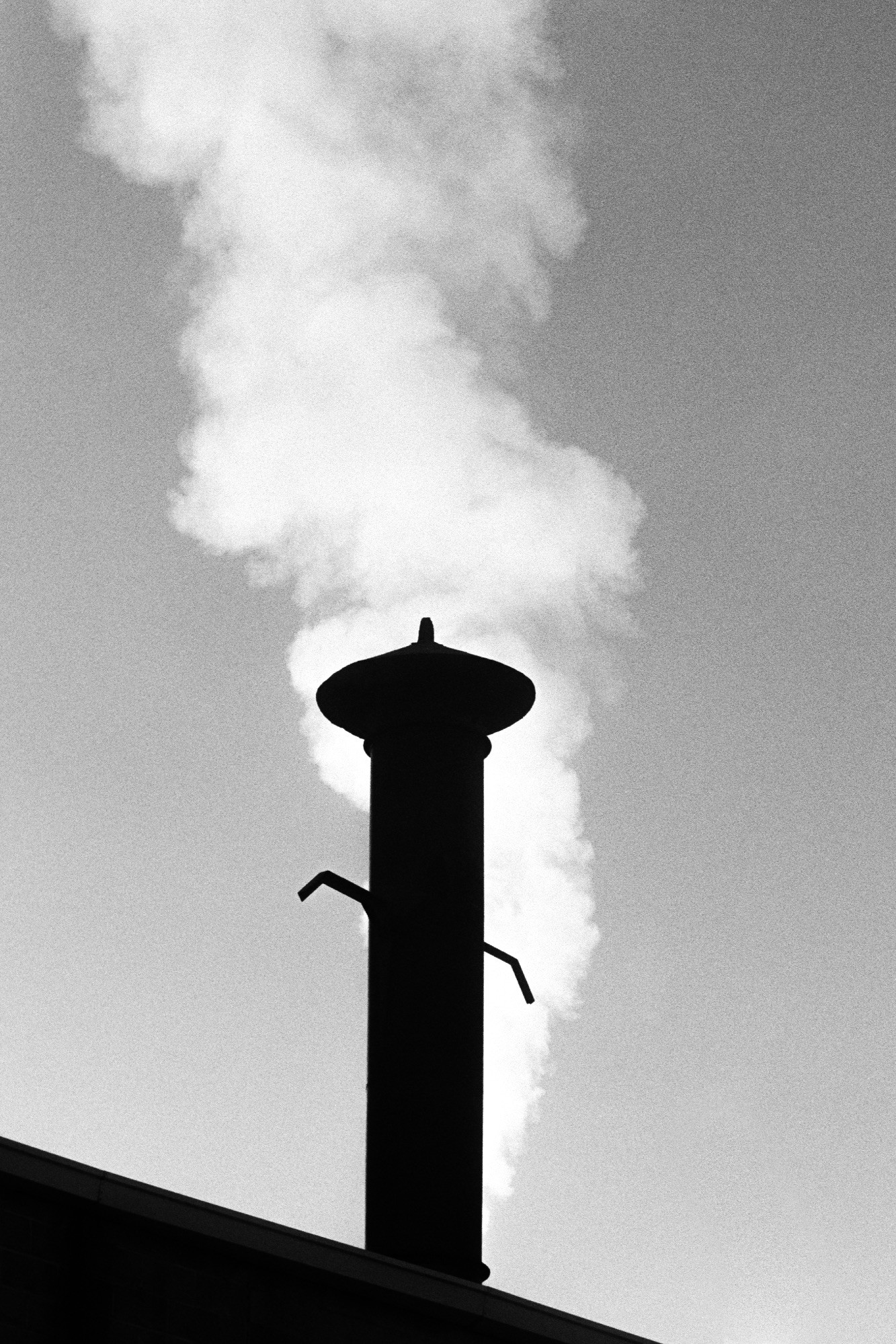 A black-and-white photo of the silhouette of a vent pipe with a conical cap in front of a stream of white smoke