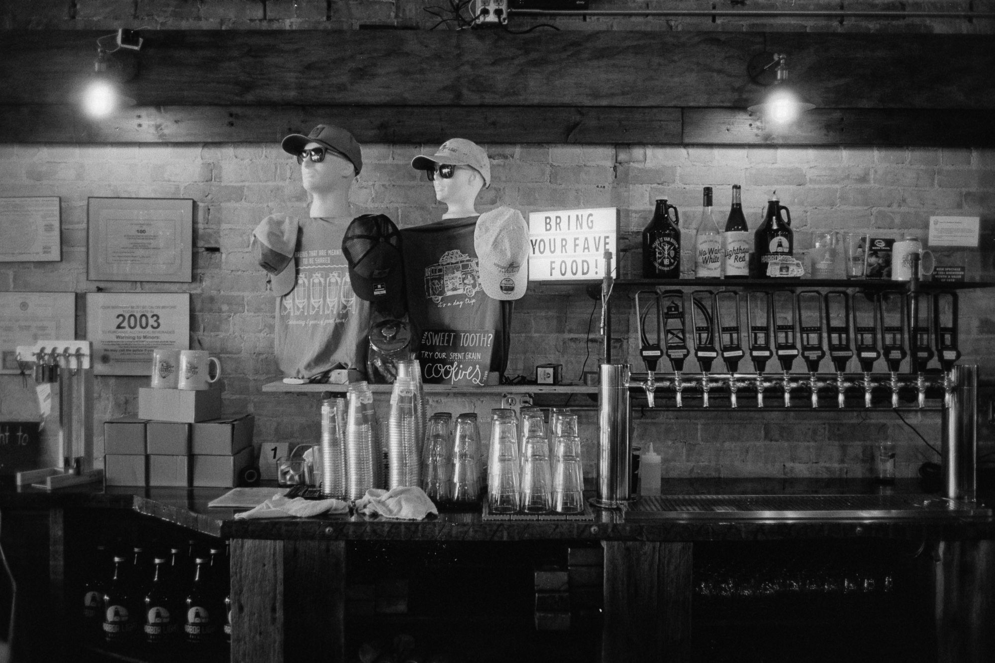 A black-and-white photo of a taproom bar with eleven tap handles, stacks of glasses, and two half-mannequins displaying store merch next to a sign reading "Bring your fave food!"