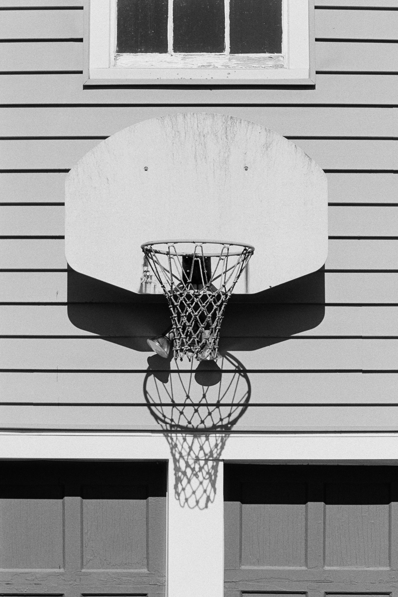 A black-and-white photo of a basketball goal mounted on the exterior of a house with the net casting a long shadow straight down into the space between twin garage doors