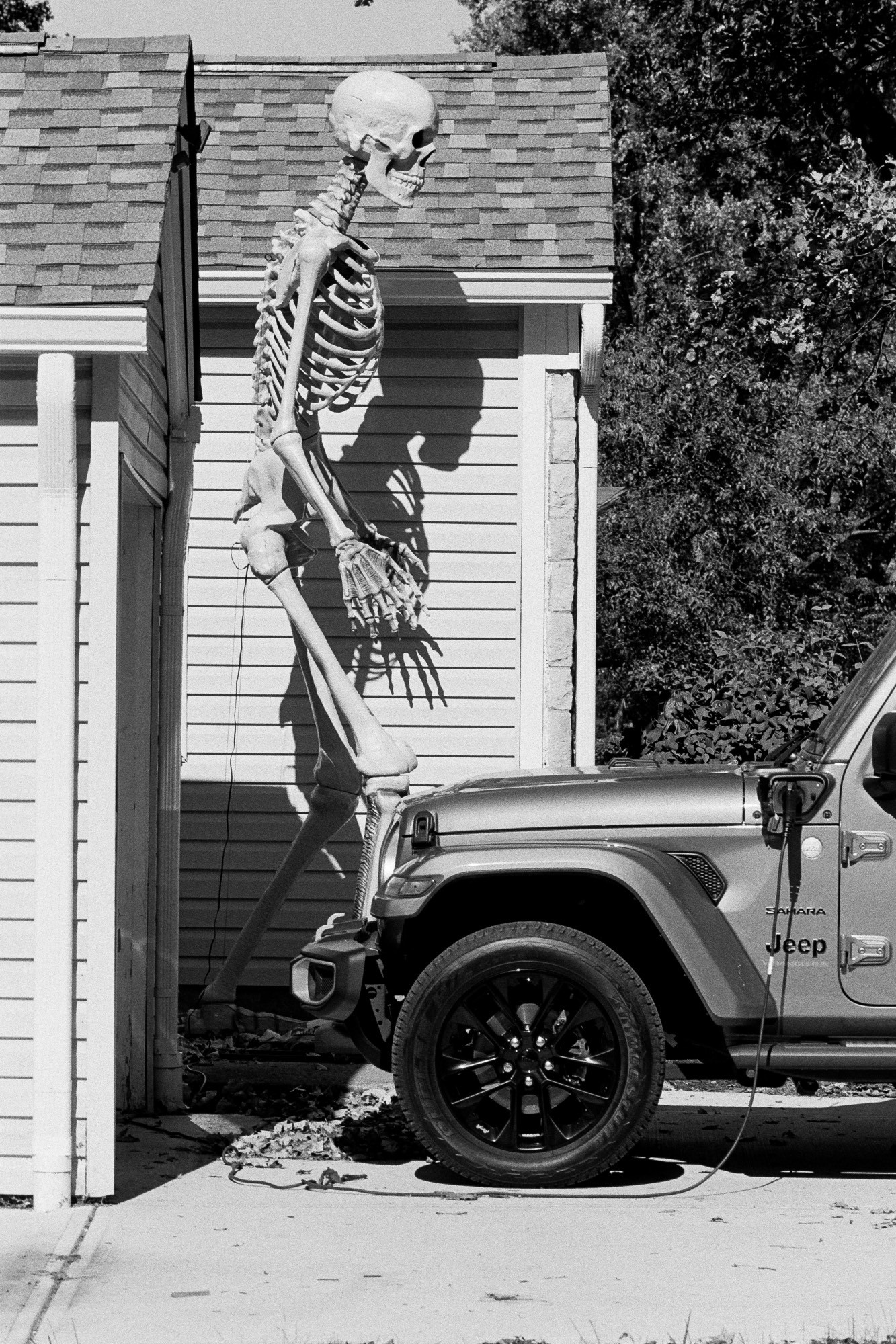 A black-and-white photo of a twelve foot skeleton decoration outside a home with an electric Jeep charging in the driveway. The skeleton is casting a dark shadow of a more squat skeleton onto the house