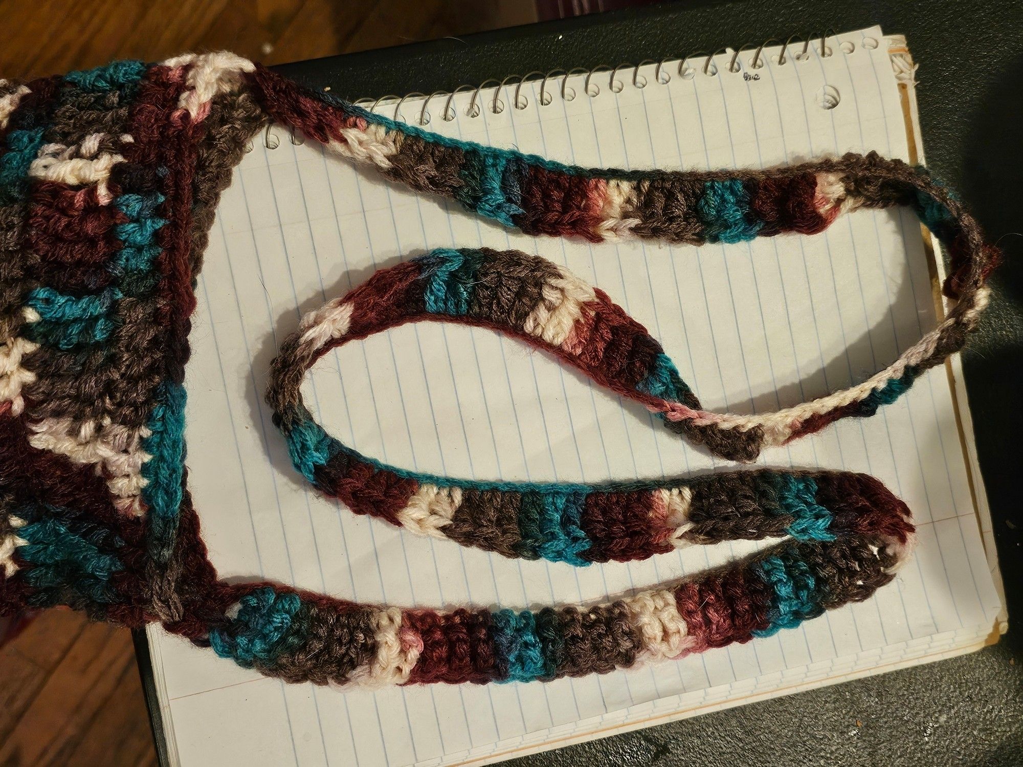 A closeup of the top of a bag and the long shoulder strap. It is made of white, purple, teal, and grey yarn.