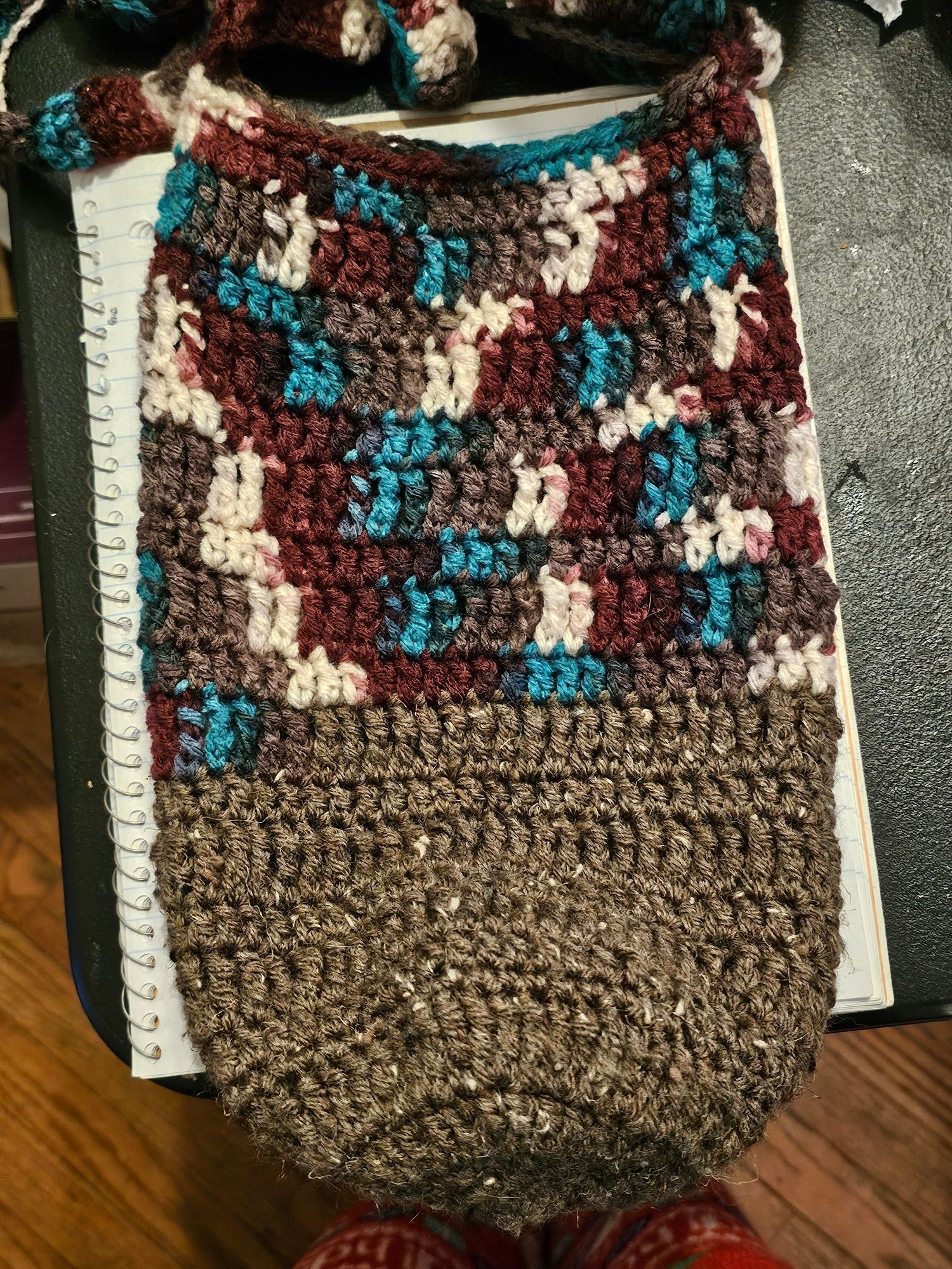 A cylindrical crocheted bag, the top half of which is white, purple, teal, and grey spotted yarn, the bottom is plain grey.