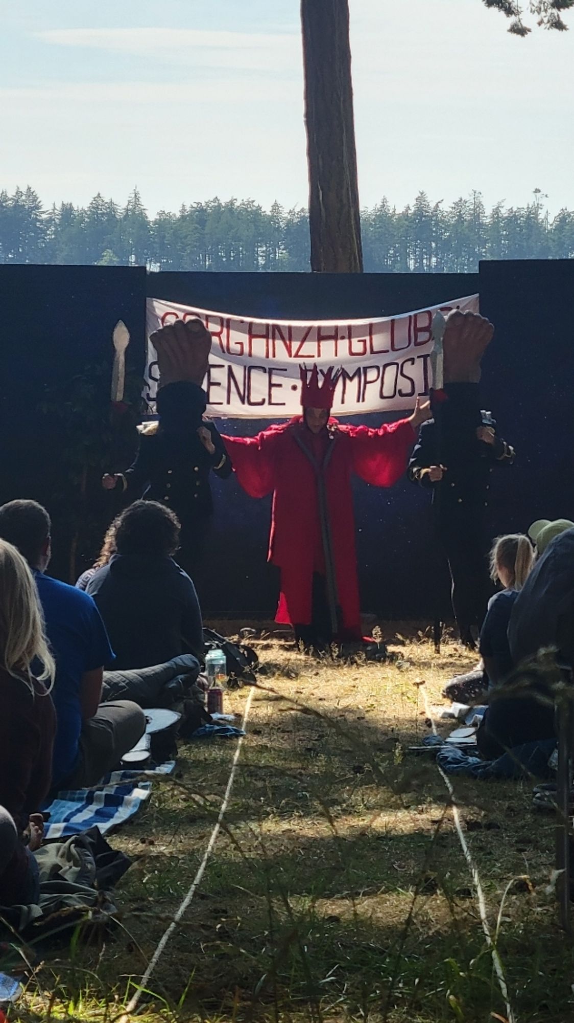 A CEO dressed as a cartoon villain with a red cape and pointy crown. They are flanked by foot soldiers who's heads are literally feet with soldier costumes. They are all in front of a 'science symposium' banner talking about a goal of mapping the universe.