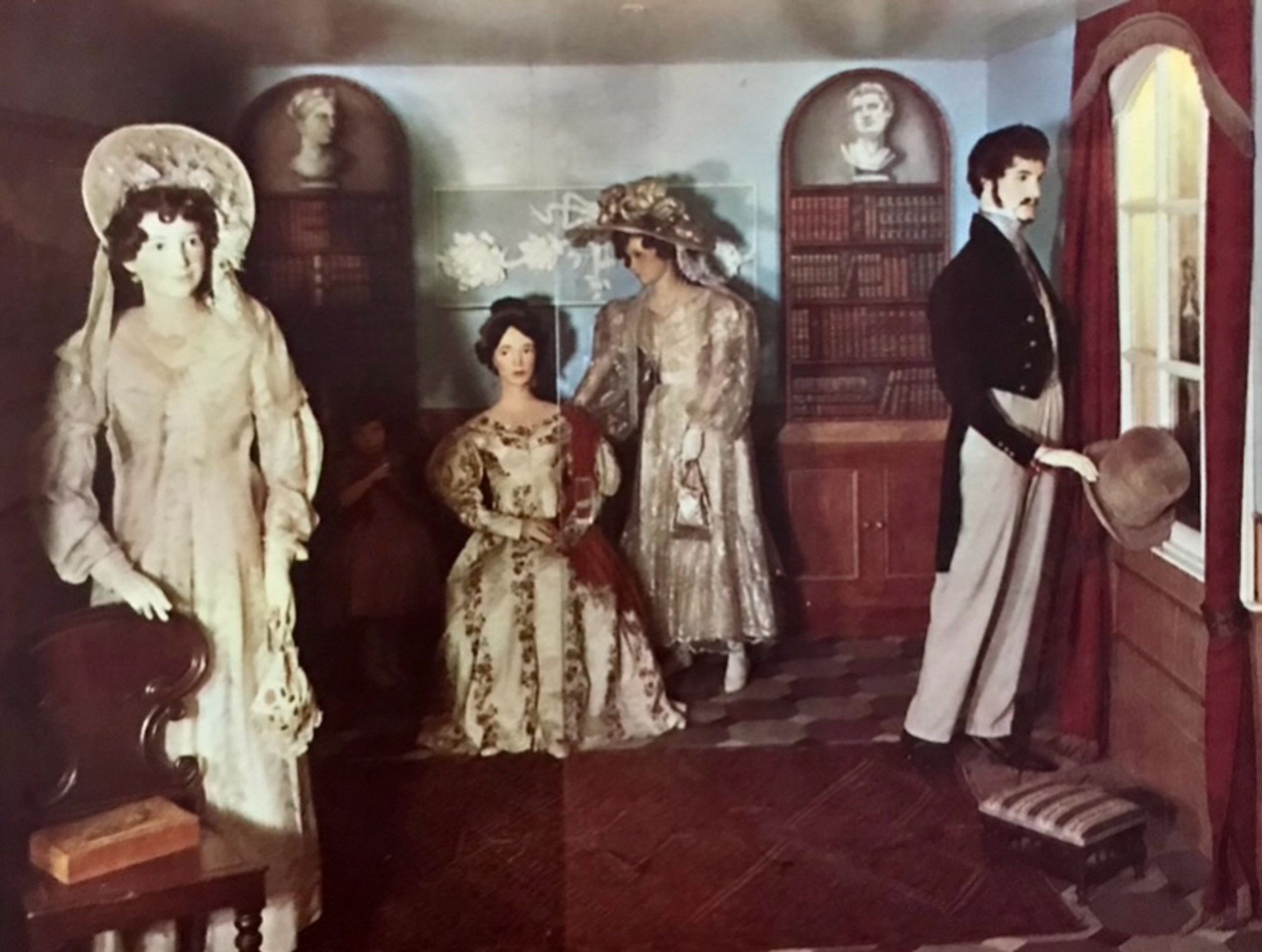 Postcard shows four creepy figures in early 19C costume, arranged in a homely library space.