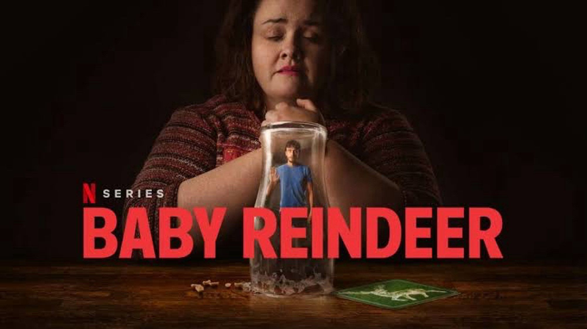 Cartaz da série “Bebê rena”, da Netflix. Imagem mostra a personagem Martha com os cotovelos apoiados sobre uma mesa. Em
cima da mesa está do personagem Donny, como uma miniatura, preso embaixo de um copo, enquanto Martha olha com angústia.