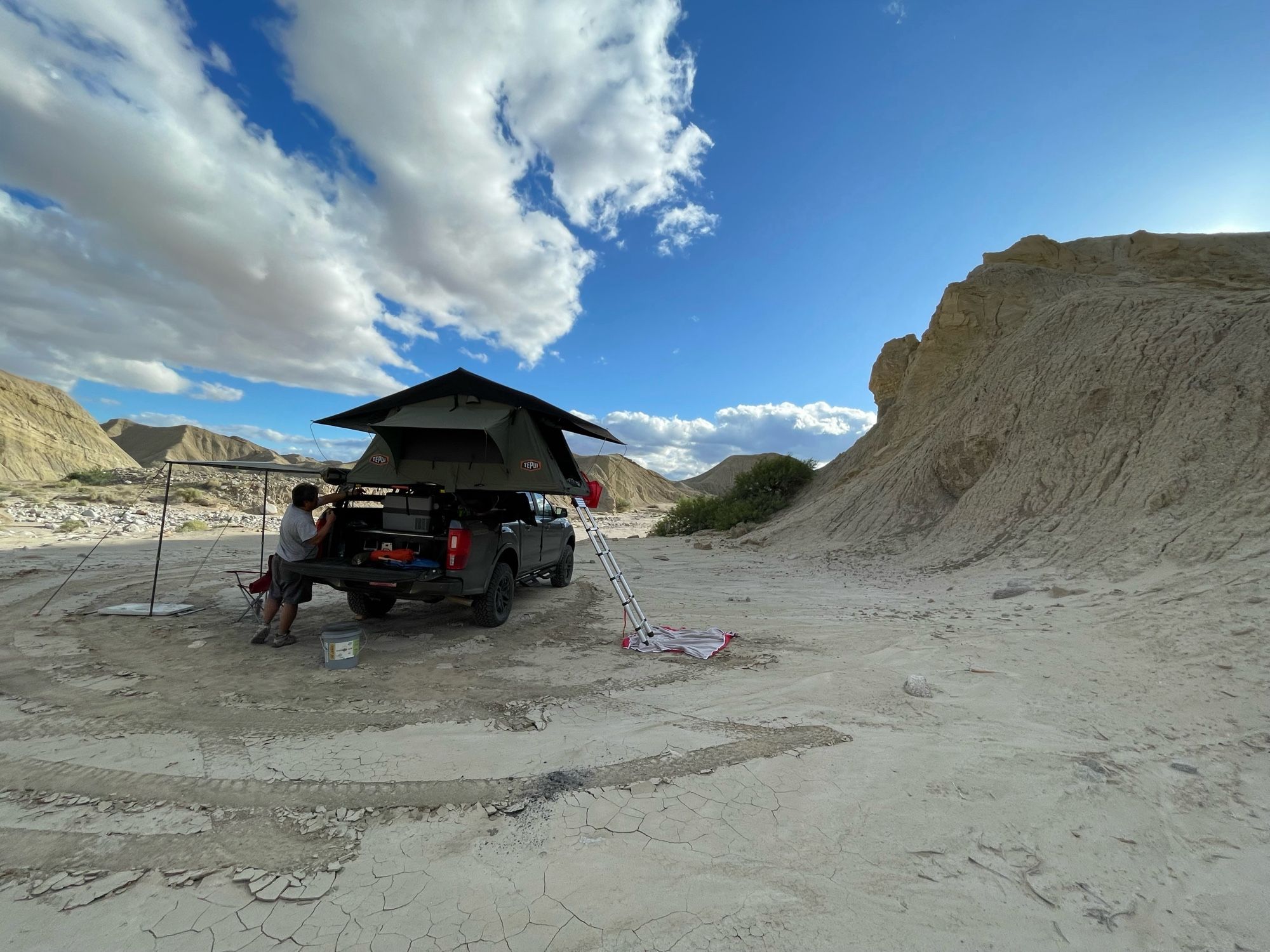 A man sets up a truck bed tent set in an area of rock and sand.