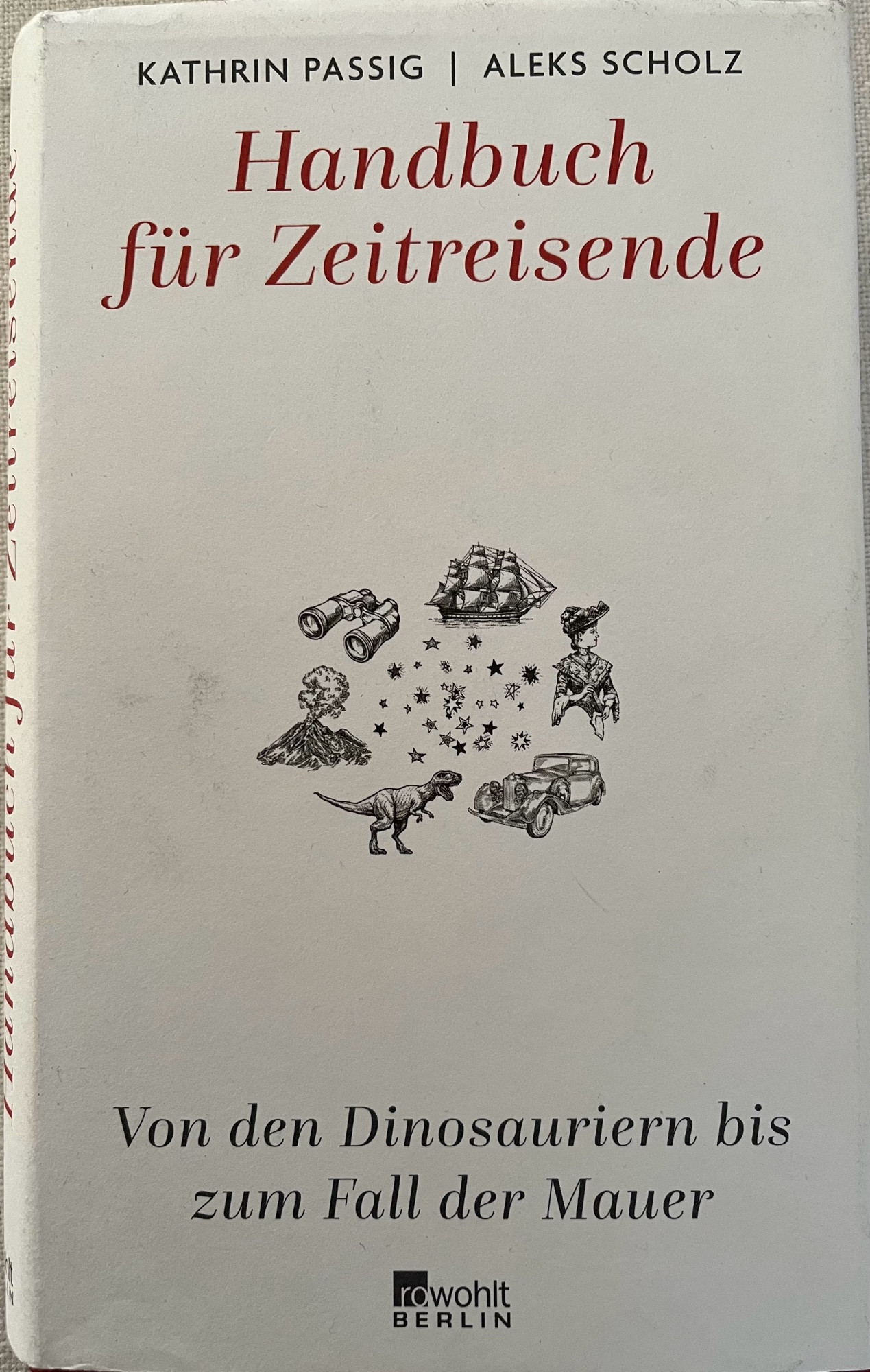 Titel des „Handbuch für Zeitreisende - Von den Dinosauriern bis zum Fall der Mauer“ von Kathrin Passig und Aleks Scholz
in der Bildmitte kreisförmig angeordnete Zeichnungen von Fernglas, Segelschiff, Frau in historischem Kostüm, Oldtimer, Dinosaurier und Vulkan sowie kleine Sternsymbole