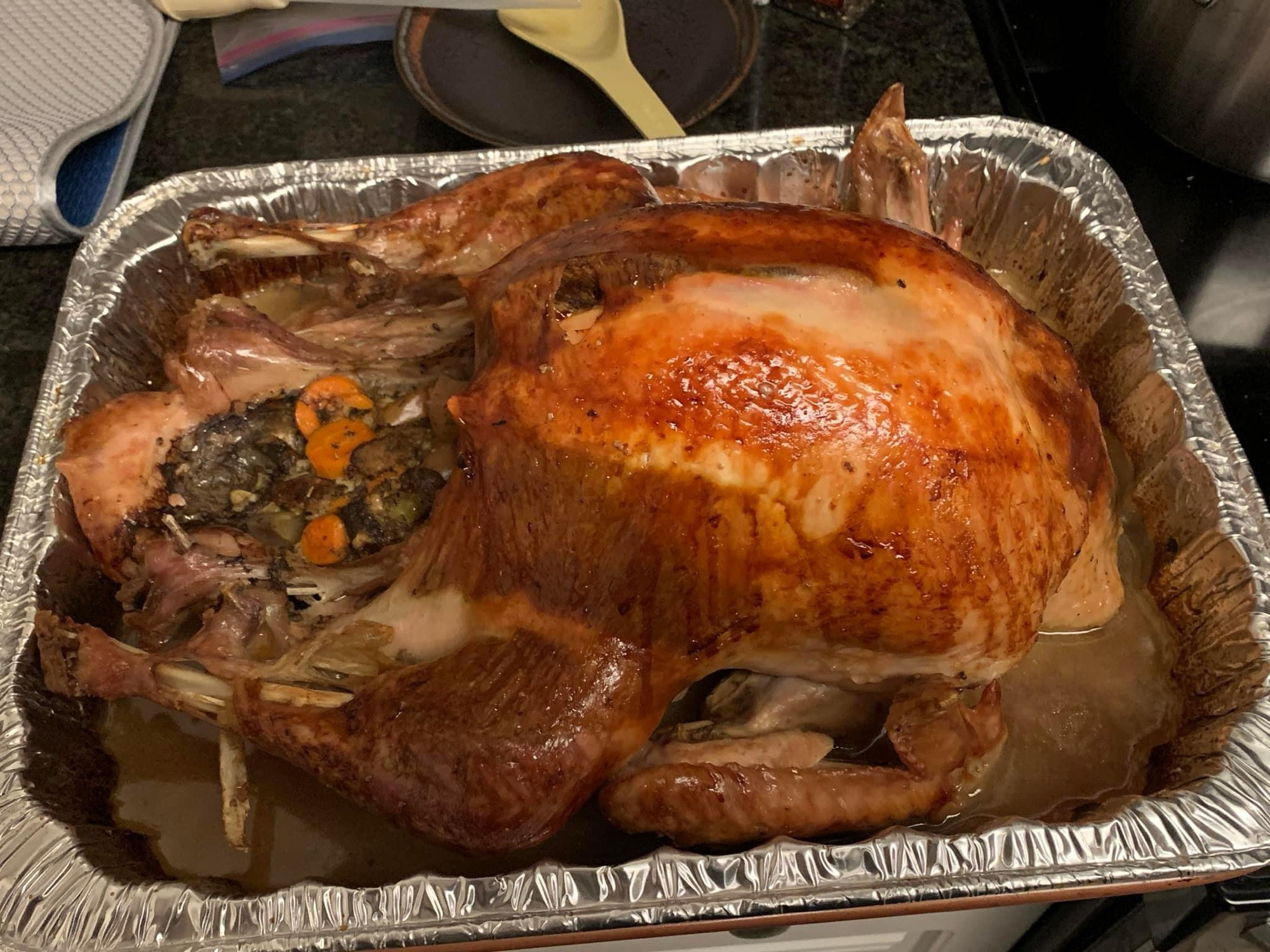 A roast turkey lounges in its own juices in a big aluminum pan with stuffing erupting out of its body cavity.