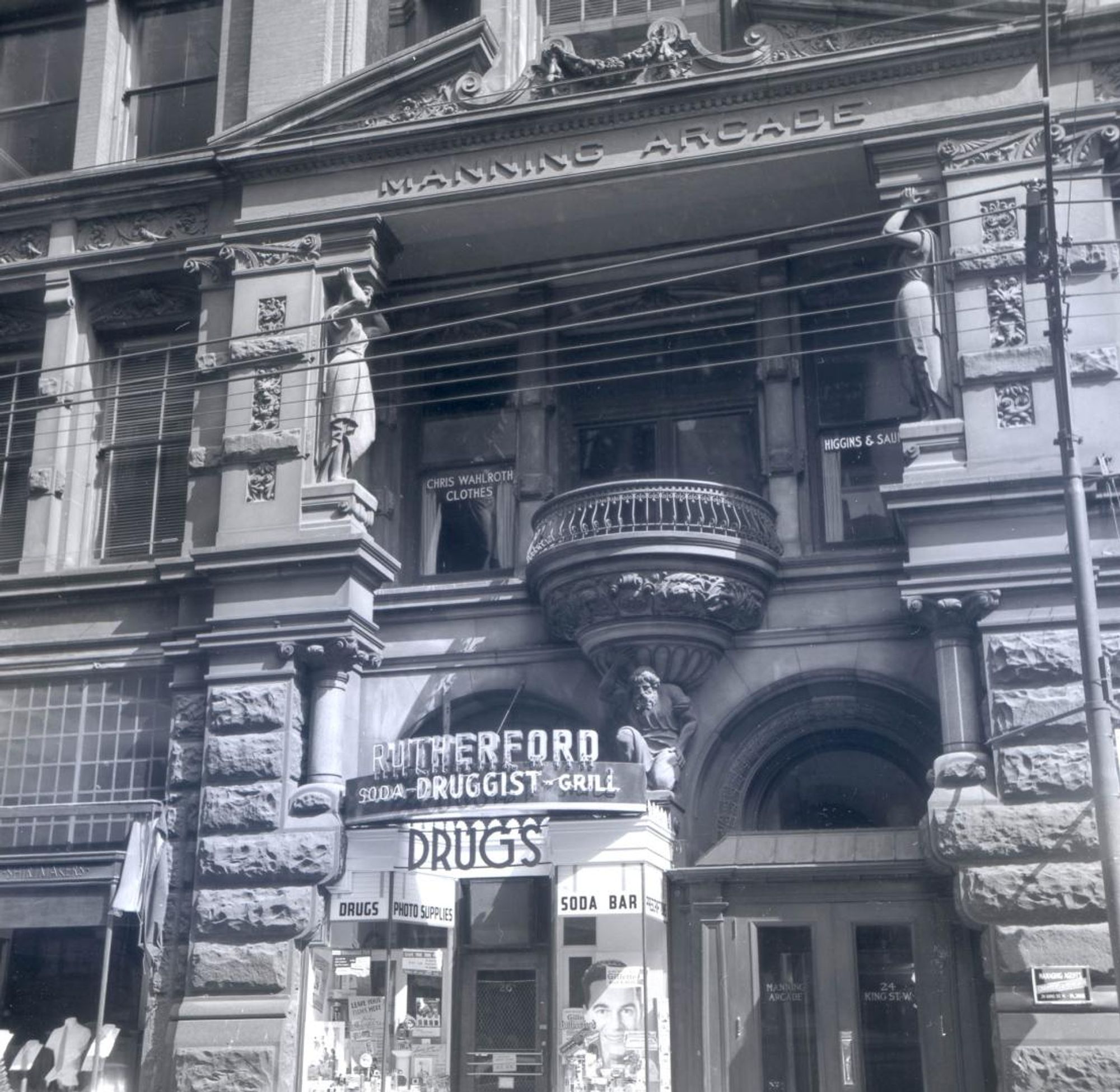 Black and white photo of an ornate building facade with a balcony, sculptures, and a soda-druggist-grill sign at street level.