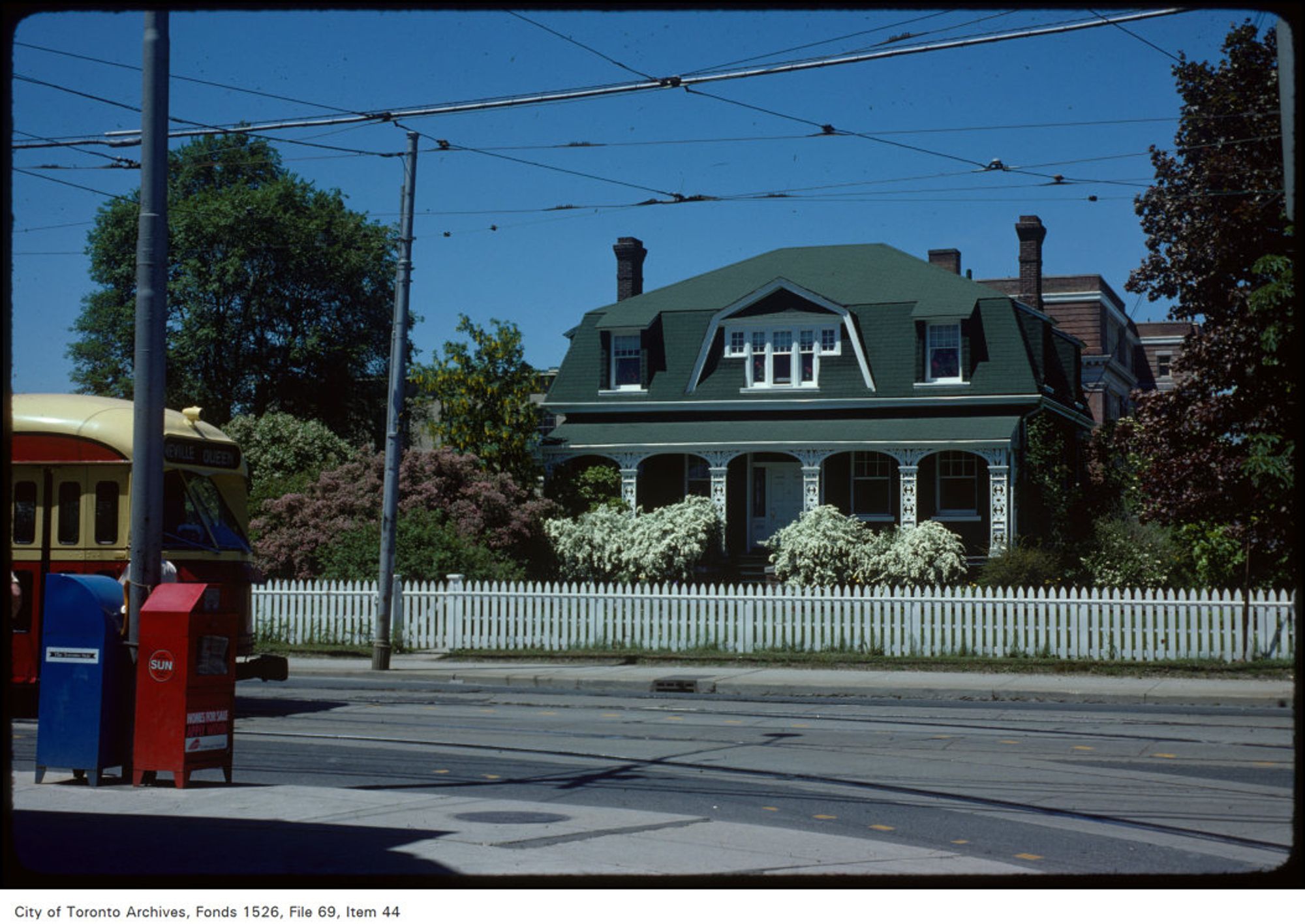 Colour photo of an old house behind a white picket fence, with a streetcar and newspaper boxes on our left.