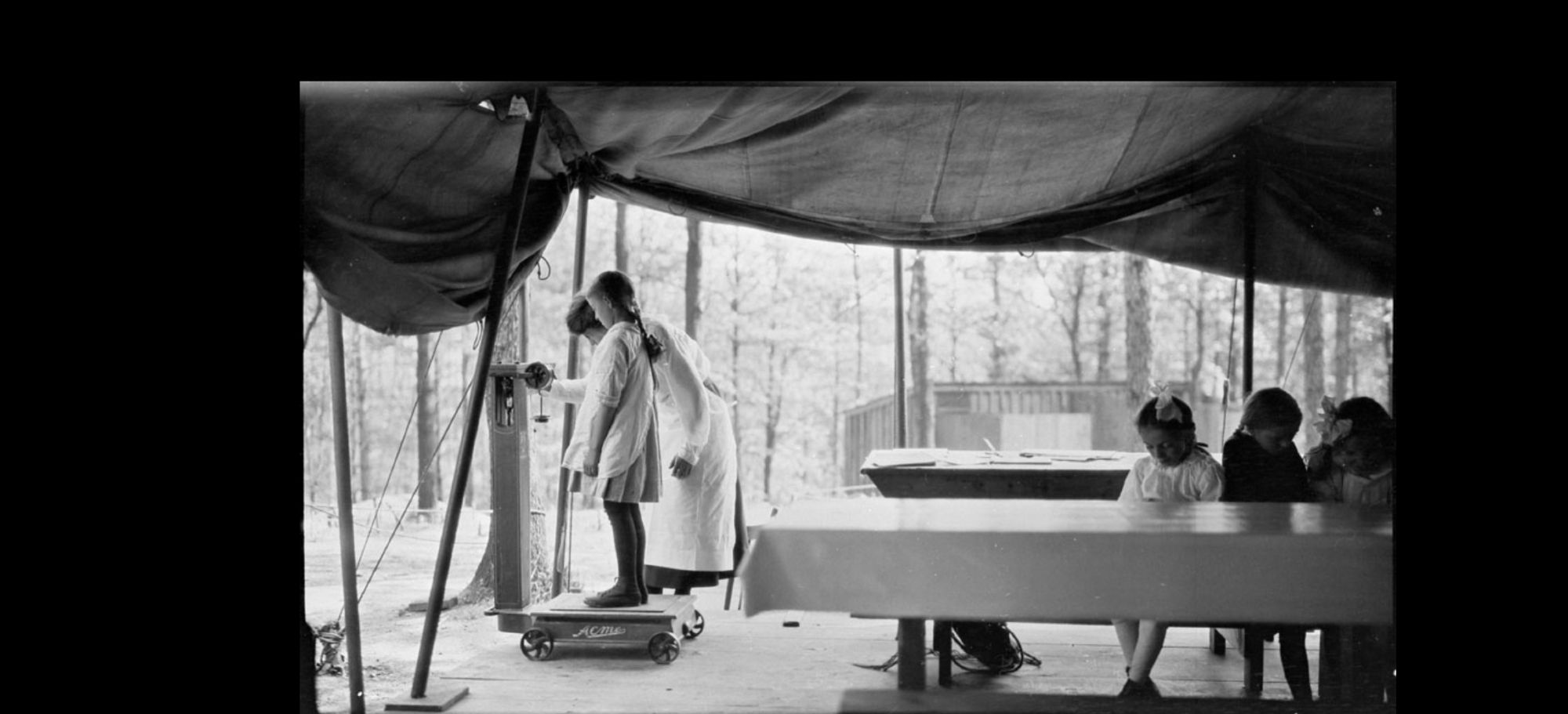 Black and white photo of a chid on a scale in a large tent, with other children waiting at a table, and trees in the background.