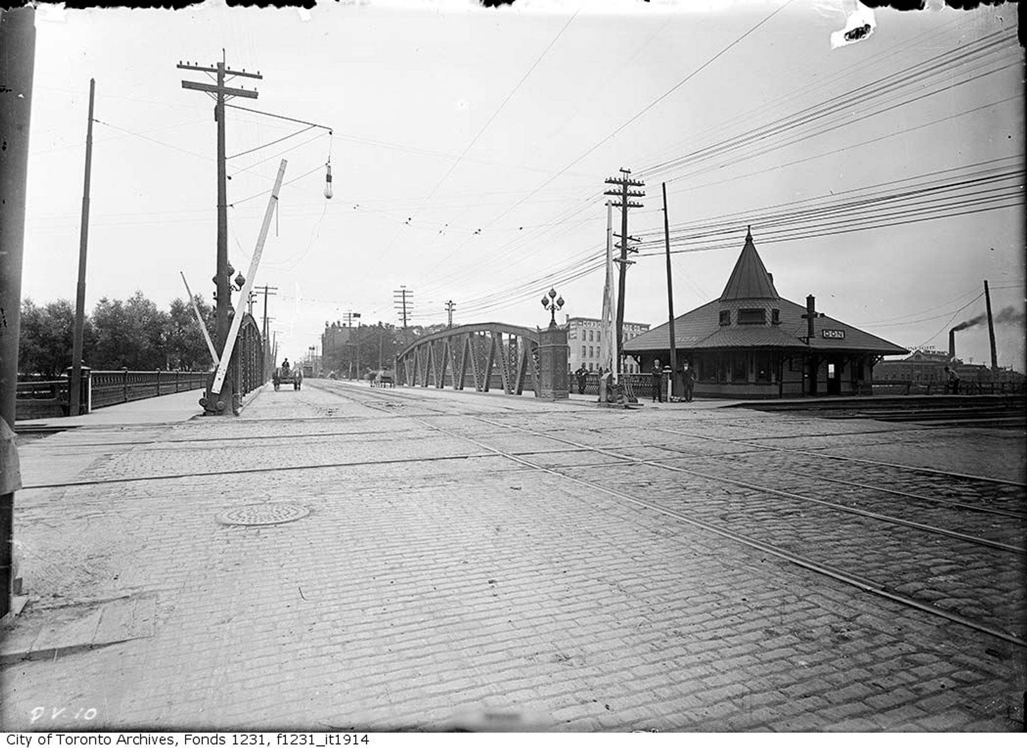 Black and white photo looking across an intersection, with railway tracks and a small train station with a bridge to our left.