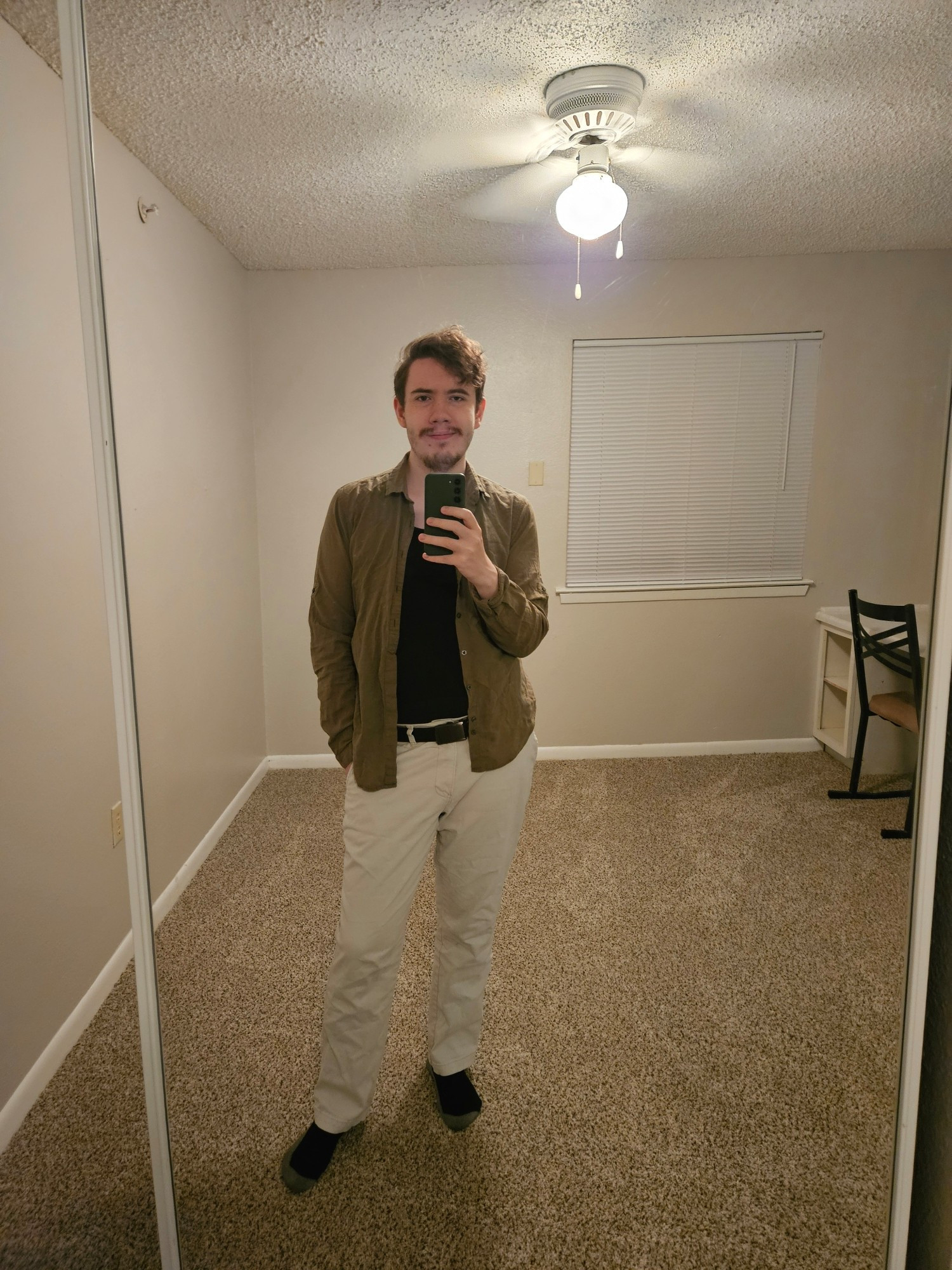 Mirror selfie taken by the same man in the same outfit in an otherwise empty room. He is looking directly at the camera and grinning slightly