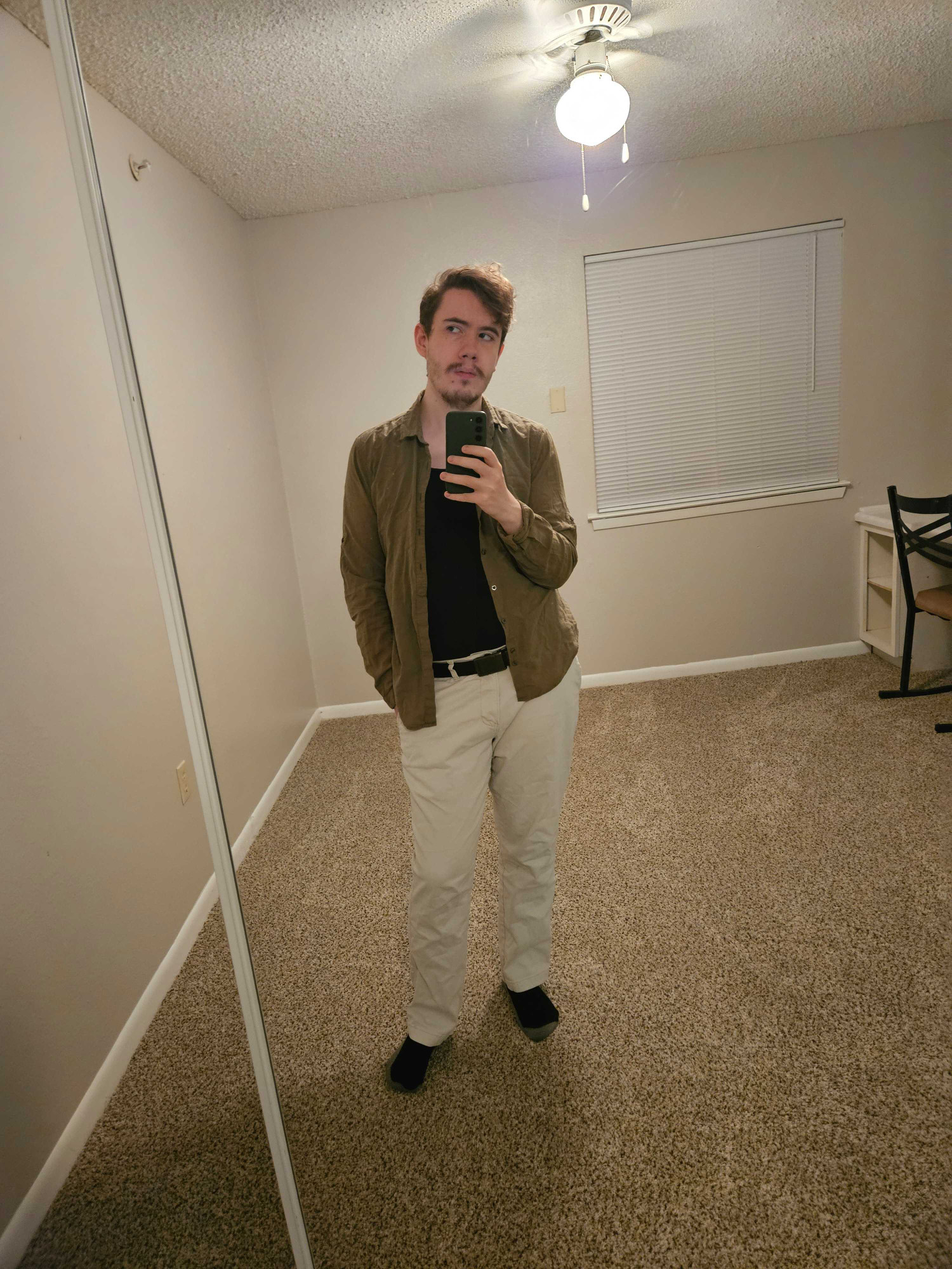 Mirror selfie taken by the same man in the same outfit in an otherwise empty room. He is facing the mirror but his eyes are off to the side.