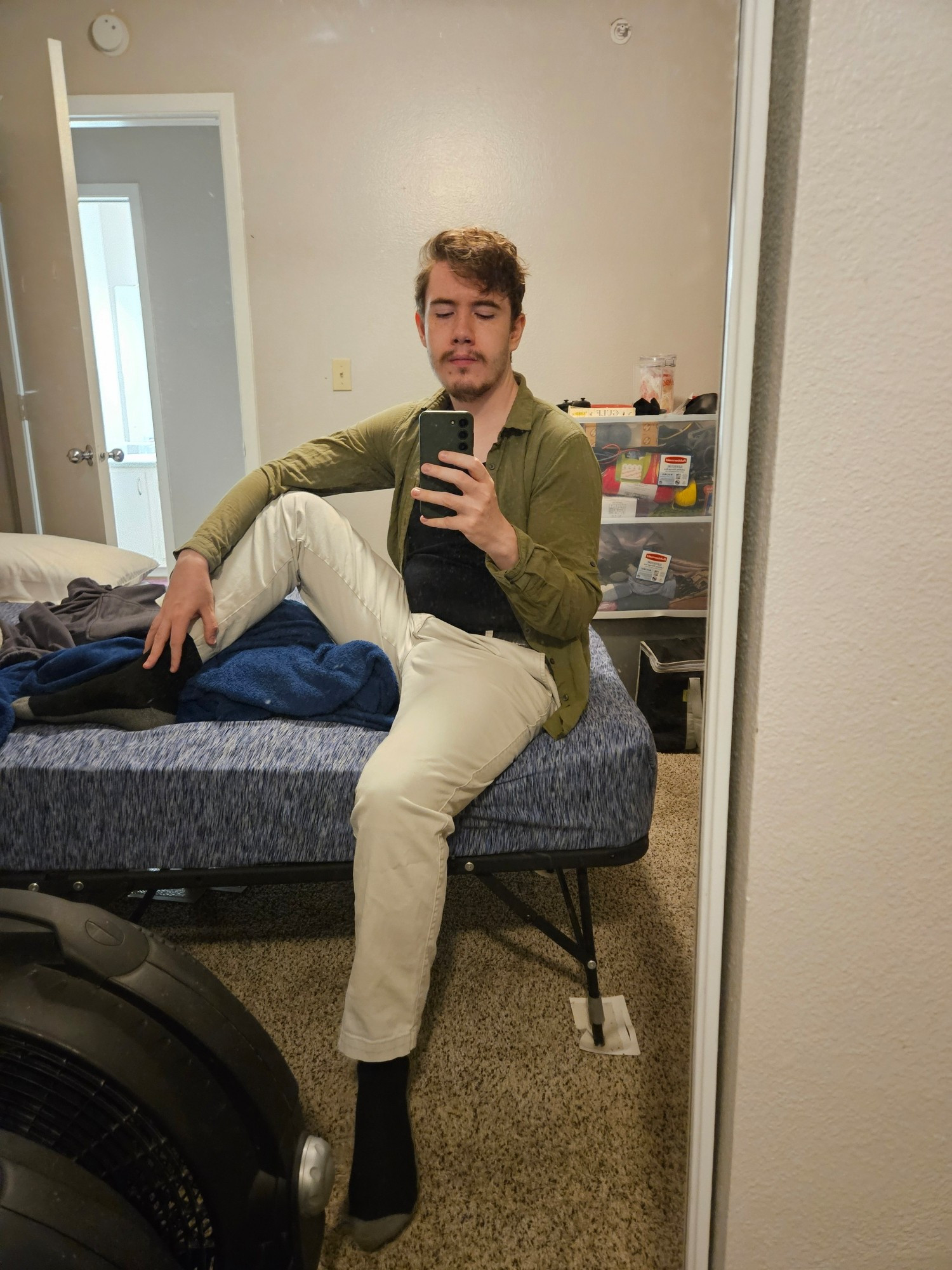 Mirror selfie taken by a man dressed in a black shirt tucked into white pants with an open green long-sleeved button-down shirt over it all. He is sitting on a bed with one leg propped up. The room is a disaster.