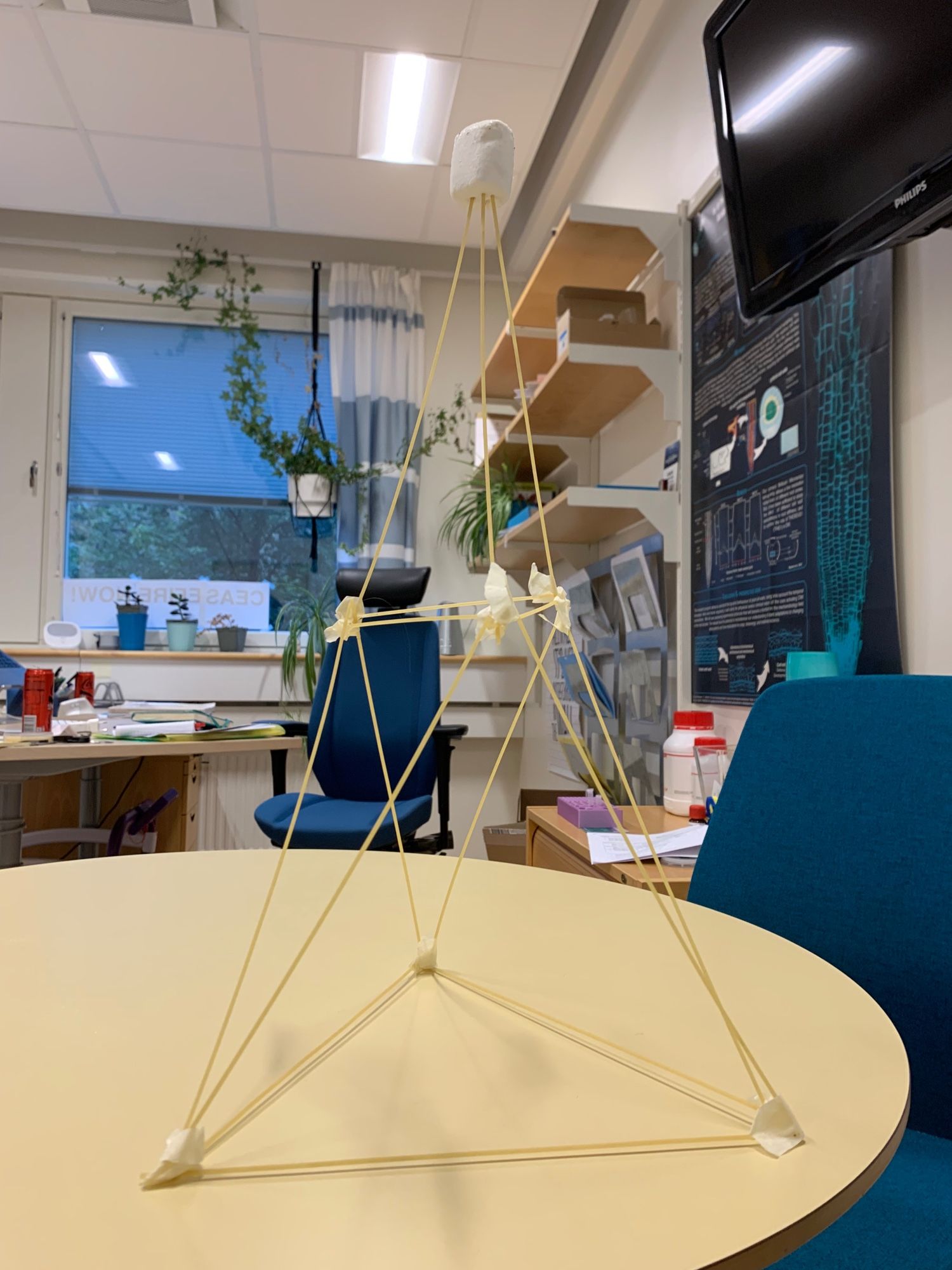 A picture of a tall triangular tower made from uncooked spaghetti sticks, held together with tape, and topped with a marshmallow. The tower is sitting on a table in an office. This structure is part of the spaghetti challenge, where participants build the tallest freestanding tower they can using only spaghetti and tape to support a marshmallow at the top. My team didn't win, but like me, this structure may not be the best, yet it stands strong and persistent.