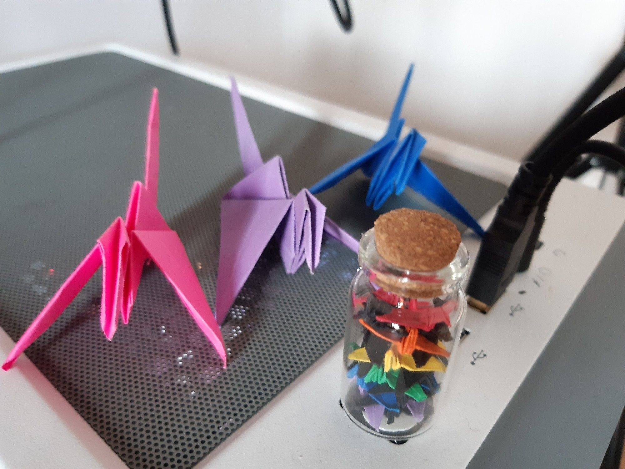 a small corked bottle of rainbow coloured and black origami paper cranes attached to the power button of a white pc case, behind it are three much larger paper cranes making up the colours of the bisexual pride flag