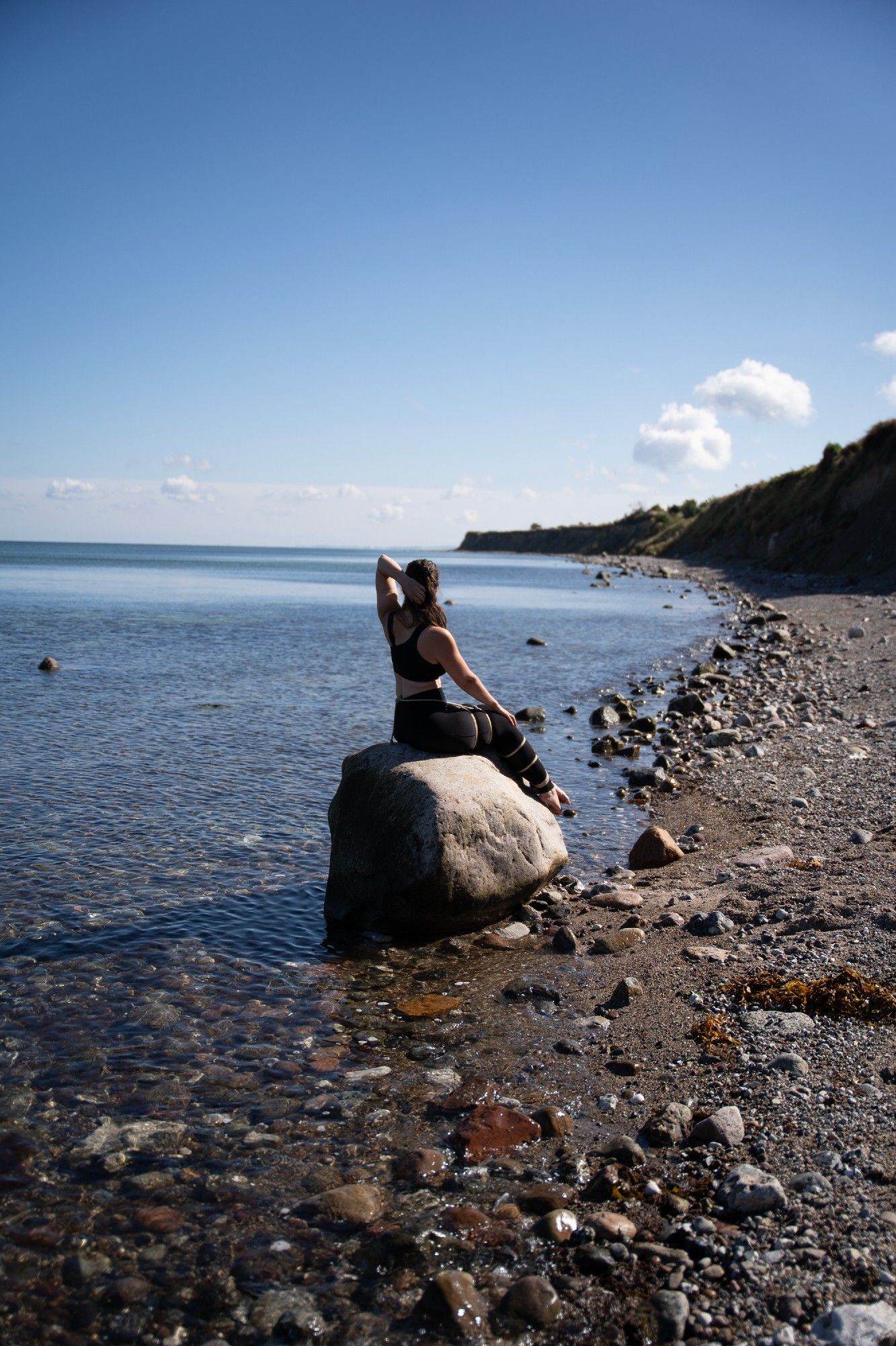 A women sitting on a stone, them placed at the shore of the sea, in a pose like the mermaid from Copenhagen. Her legs are tied together. in the background you can see the ocean and the coast. the sun is shining.