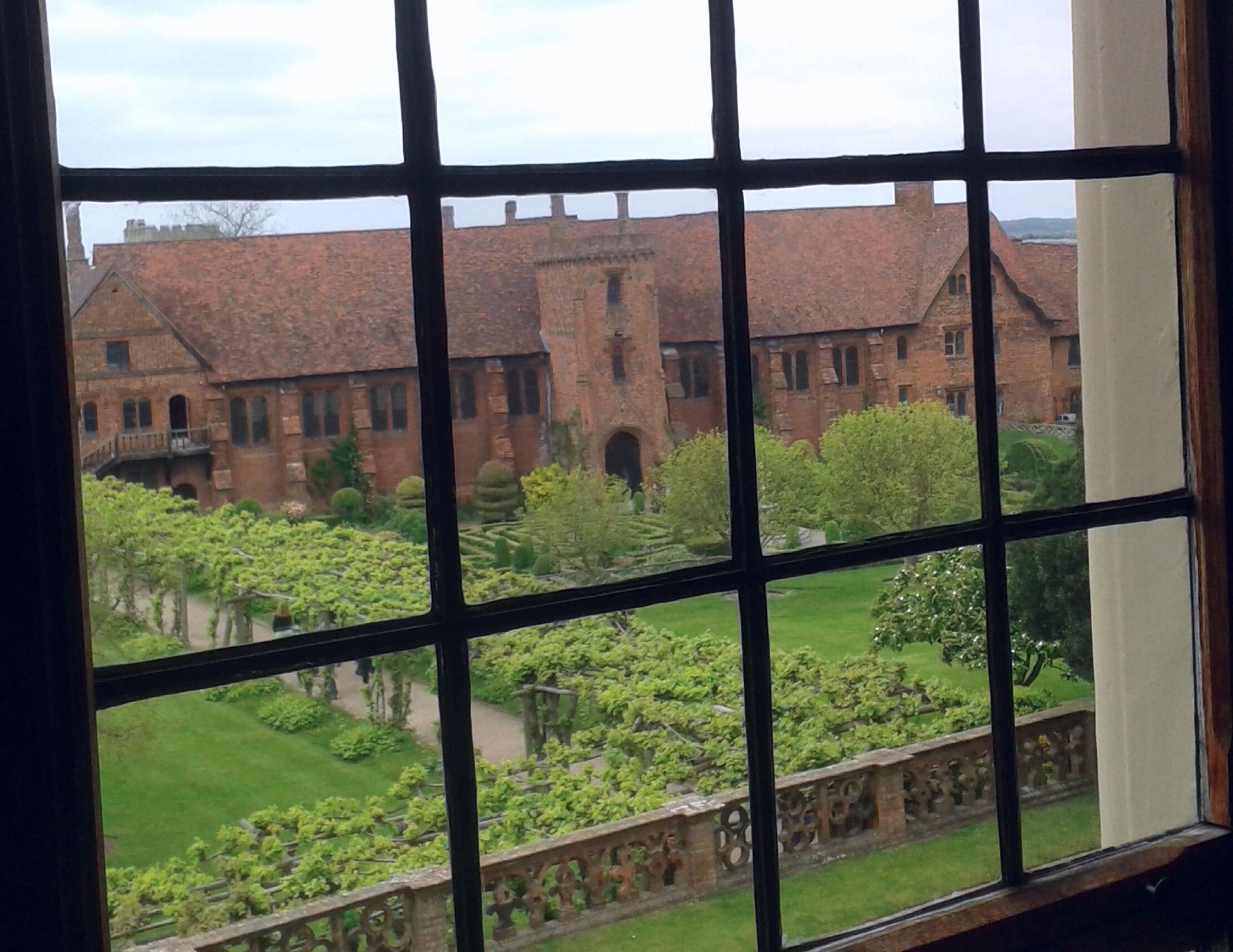 The full width of the remaining orange brick Tudor bulding of the Old Palace at Hatfield can be seen across the beautiful gardens with rows of small trees. All this seen through a barred window in the foreground in the "new" Hatfield House