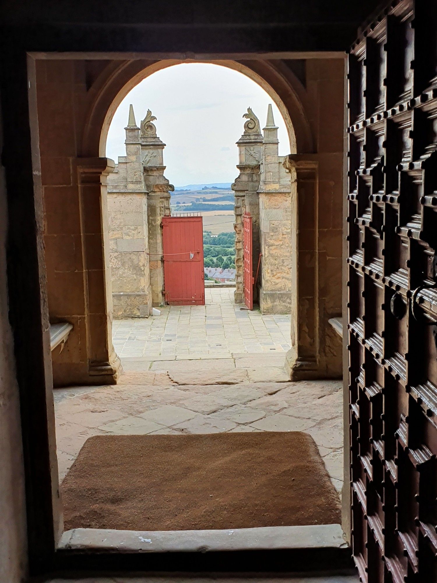 View through a heavy old decorated wooden door which is open, out through an outer arch and through a big red outer gate, with one gate closed and one open through which can be seen fields and trees in the far distance under an overcast sky.