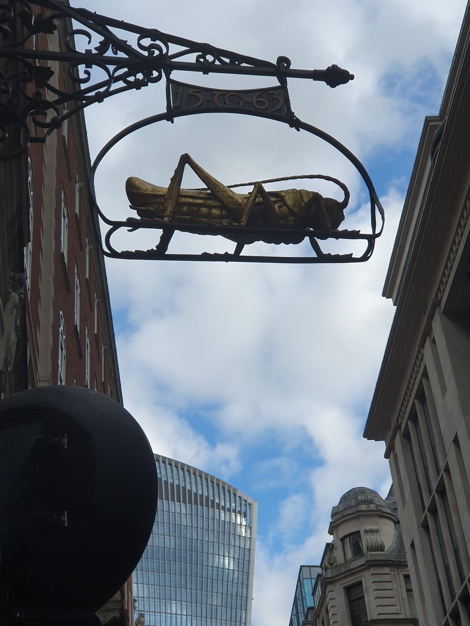 Un the foreground in the sky, the sign of the Golden Grasshopper at 68 Lombard Street in London. The initials TG can be seen above with the date 1563, referring to Elizabethan merchant Thomas Gresham who owned a bank on this site. In contrast, the ultra-modern Walkie-Talkie building can be seen in the background.