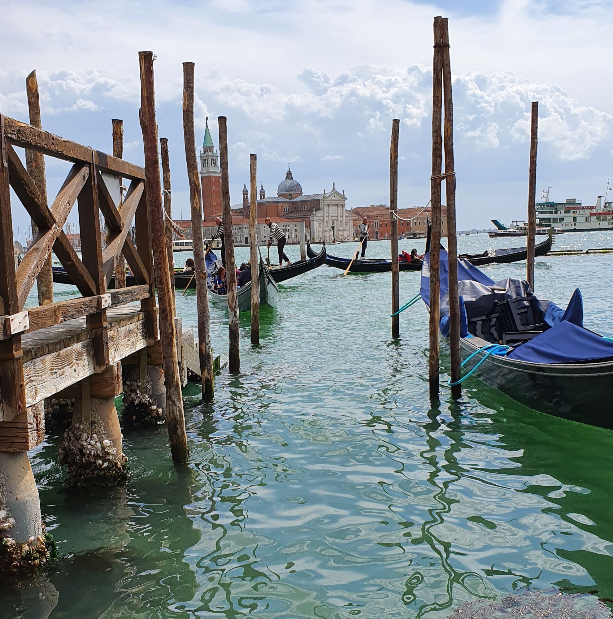 In the foreground, the murky green lagoon waters of Venice with a number of tall wooden stakes in the water, to one of which is moored a canopied gondola not in use. In the middle distance can be seen three gondolas, two of whose high bows appear to be just touching as the gondoliers on each stand working their oars and wearing their traditional striped jersies. In the far distance across the lagoon, between the wooden stakes, can be seen the Palladian facade and campanile of the church of San Giorgio Maggiore on the small island of the same name. The sky above is filled with white clouds.